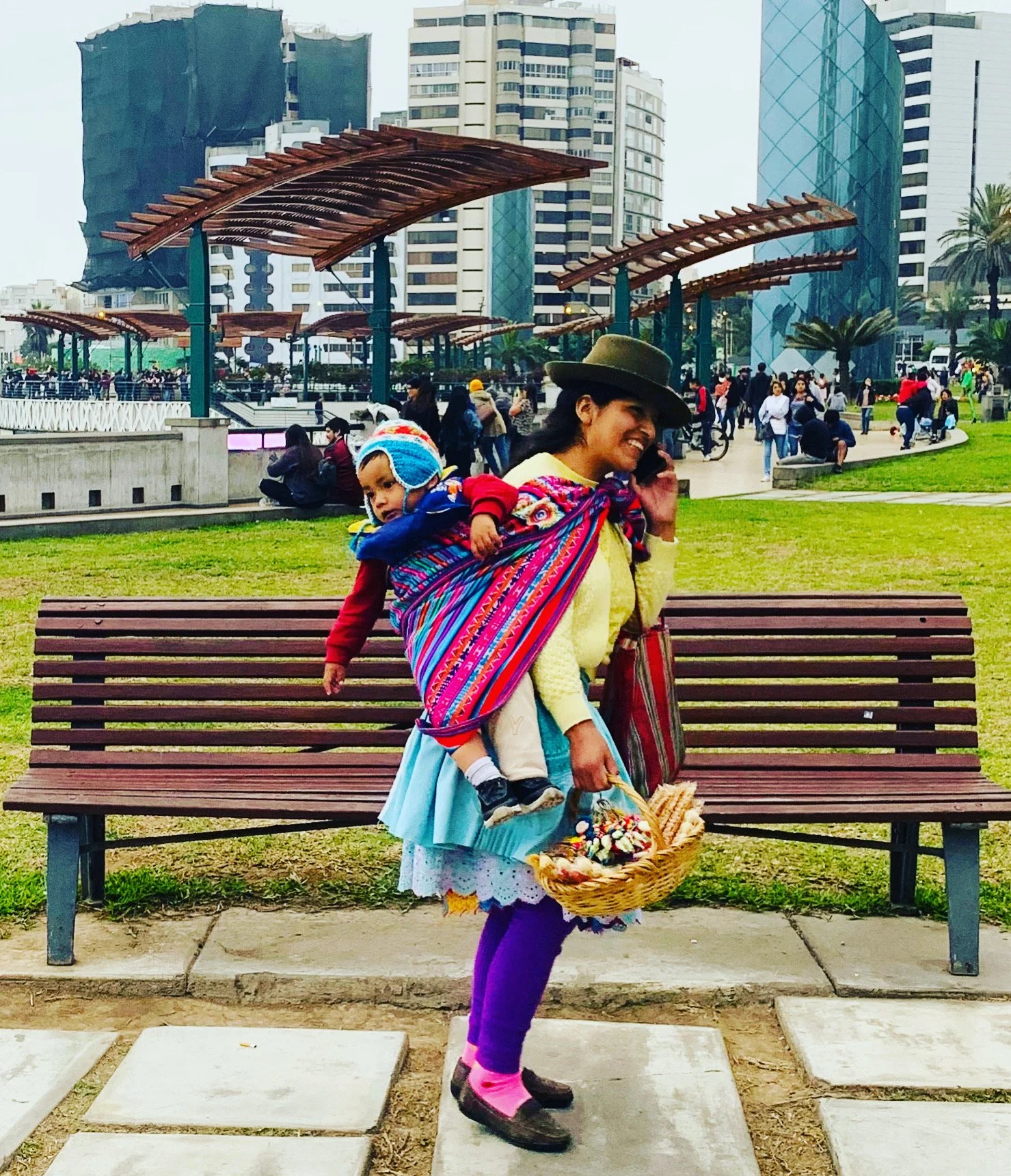 A woman walking on a city sidewalk carrying a child on her back with a colorful woven cloth. The woman is talking on her phone and holding a basket of small decorative items. The child is wearing a blue and white hat and is leaning on the woman's bac