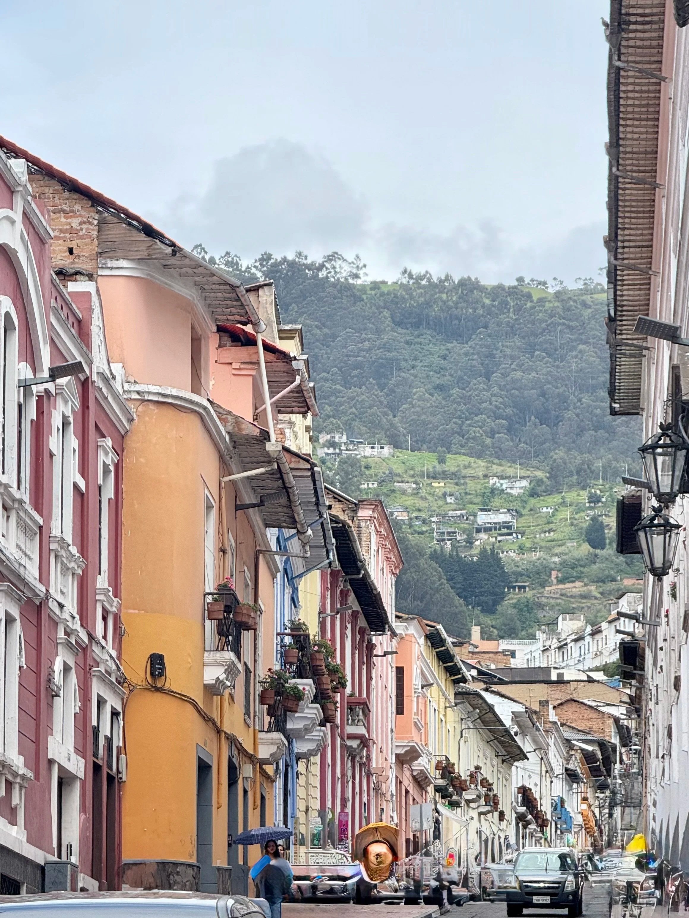 Colorful buildings on a narrow street in a hillside town with cars and pedestrians, mountains in the background on a cloudy day.