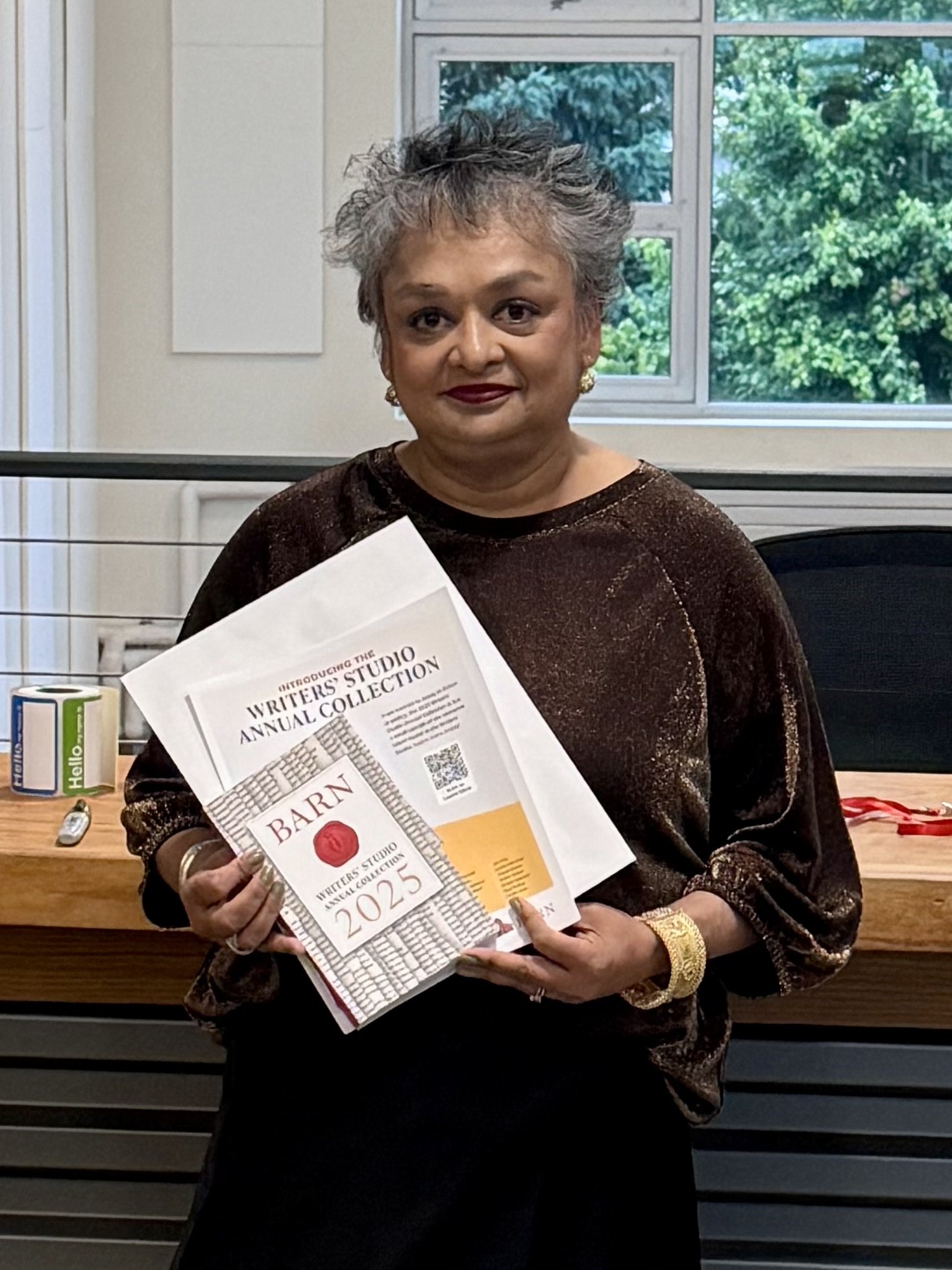 A woman with short gray hair, dark red lipstick, and gold earrings and bracelet, standing indoors near a window with green trees outside. She is wearing a brown, glittery top and holding a 2023 Writers Studio Annual Collection magazine.
