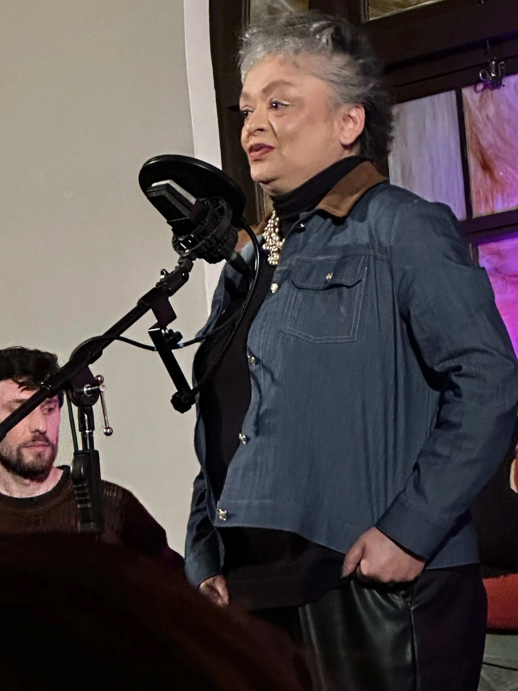 An older person with grey, styled hair standing in front of a microphone, wearing a dark jacket and a pearl necklace.