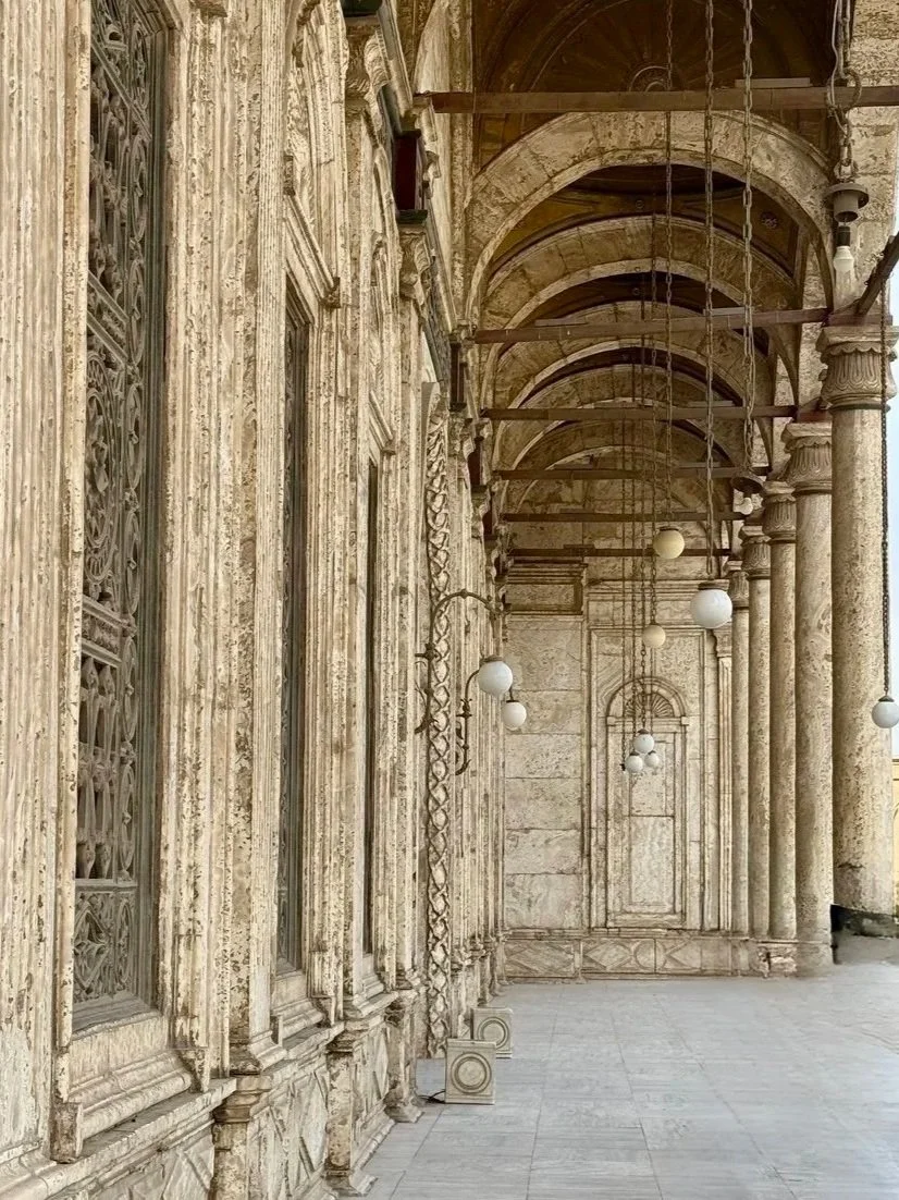 A historical building exterior with ornate stone columns and arches, decorative windows with iron grille work, hanging lanterns, and classical architectural details.