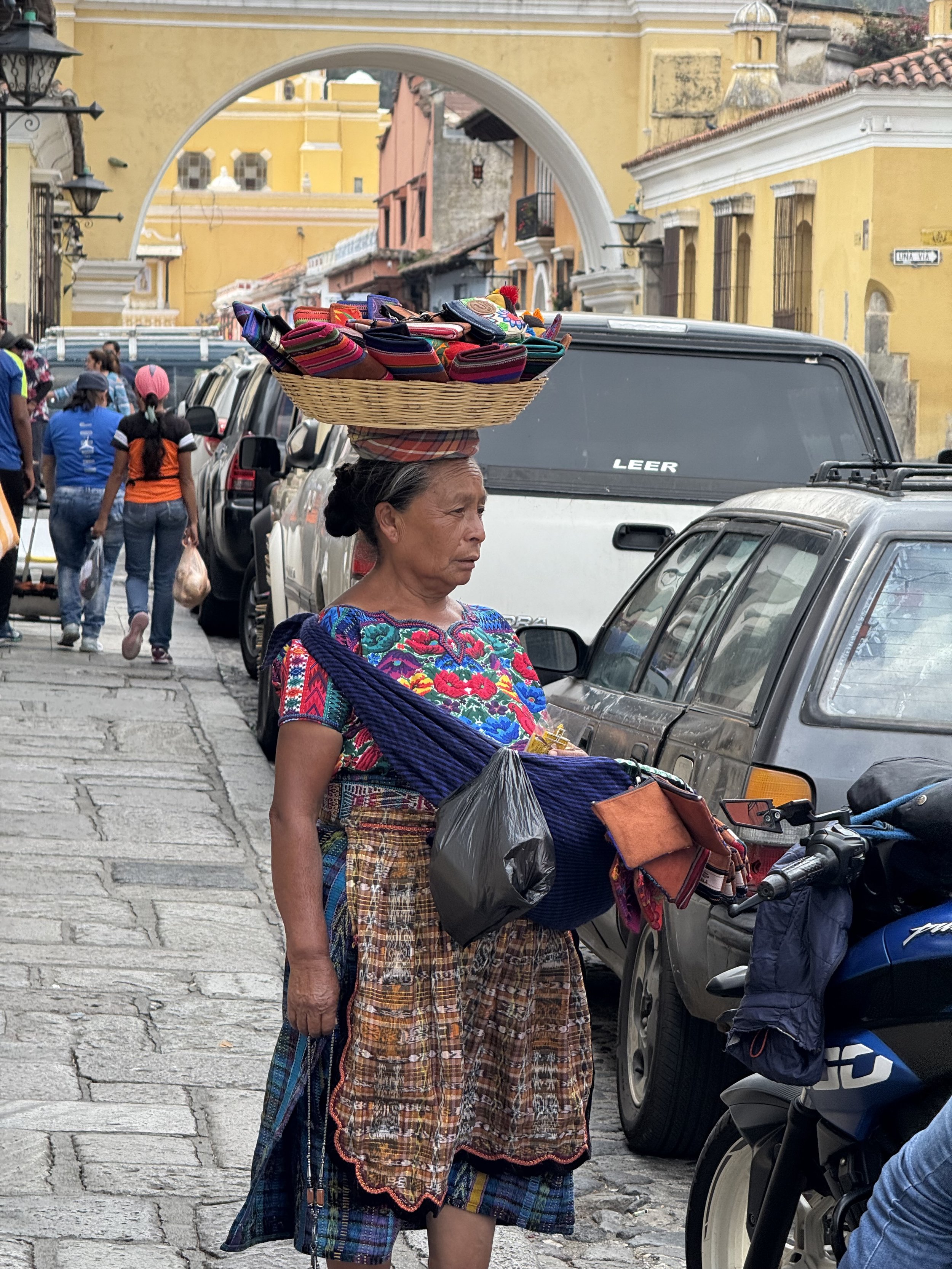 A woman with traditional clothing balancing a basket of colorful textiles on her head, walking on a cobblestone street in a historic area with yellow and pink buildings.