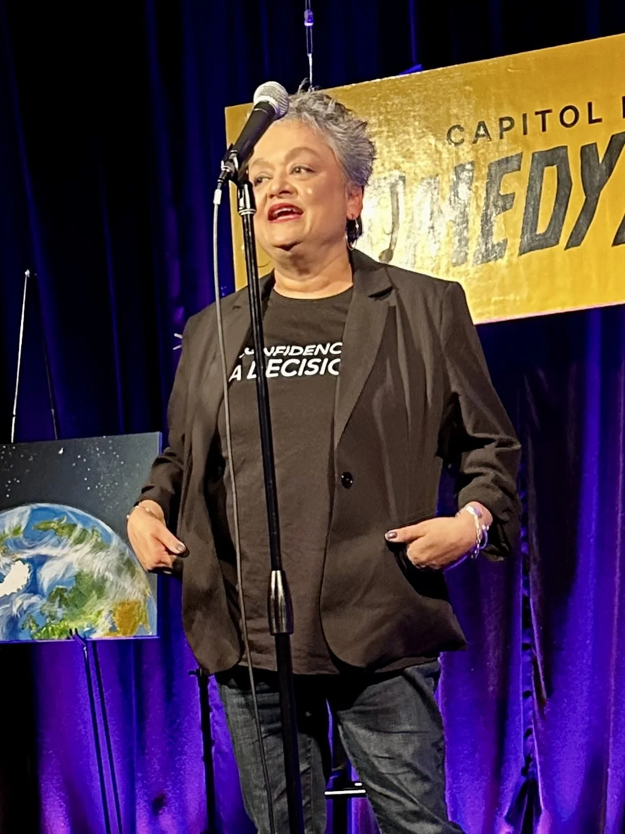A woman with short gray hair speaking at a podium on a stage, wearing a black blazer and t-shirt, with a microphone in front of her and a banner in the background that reads 'Capitol.'