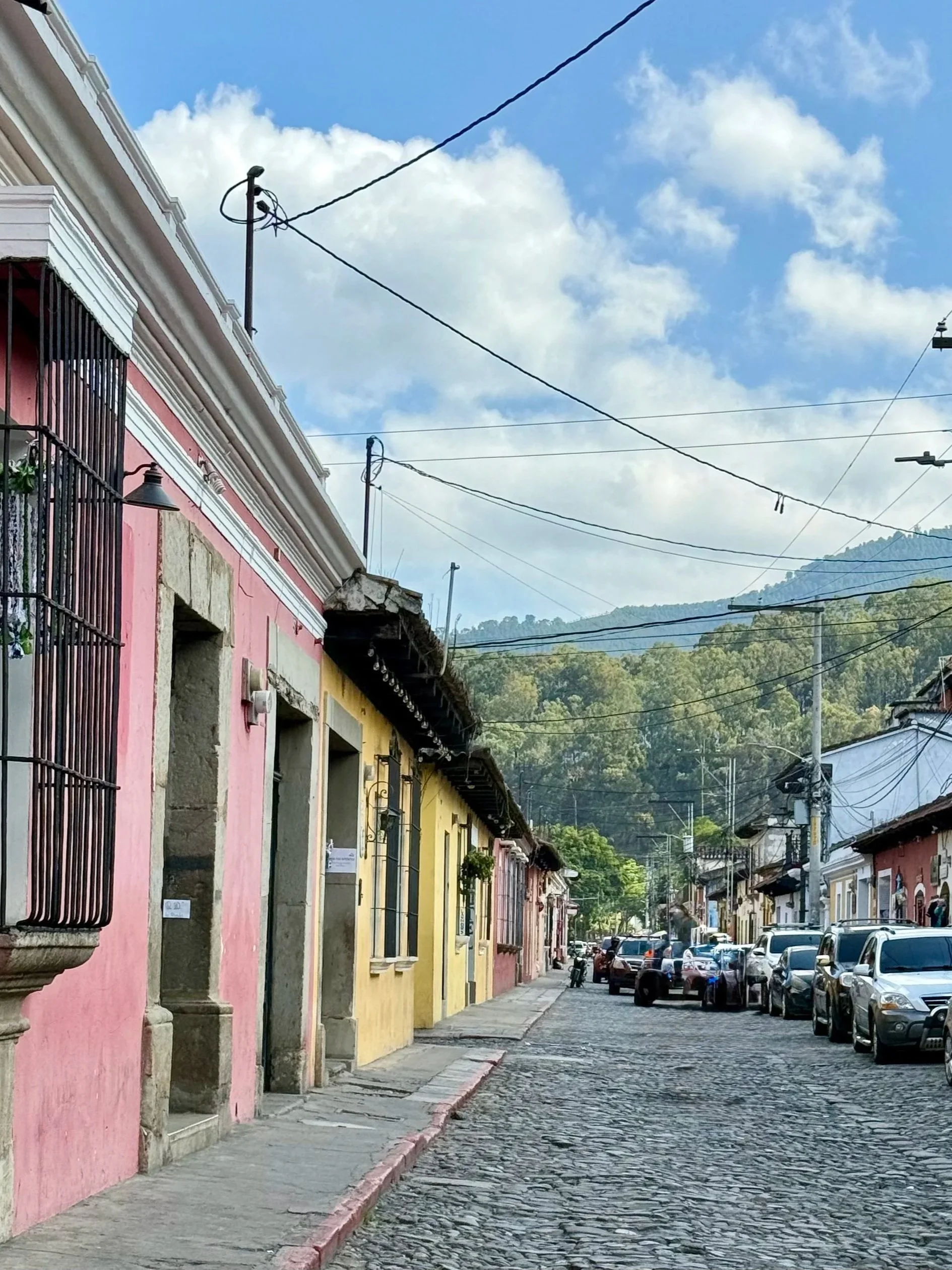 Cobblestone street lined with colorful buildings, parked cars, and utility wires, with green hills in the background under a partly cloudy sky.