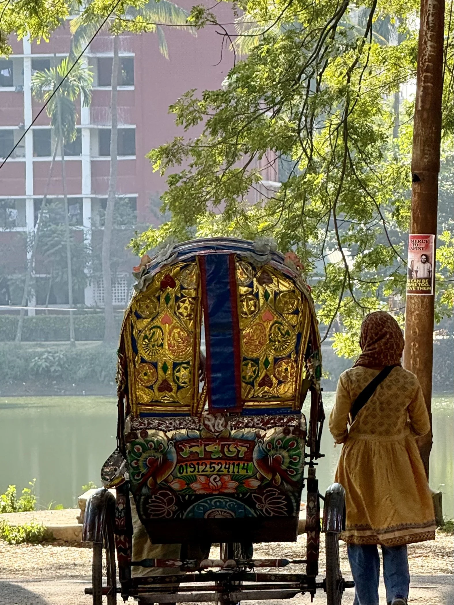 Colorfully decorated rickshaw pulled by a woman wearing a yellow coat and headscarf, standing beside a lake with trees and a building in the background.