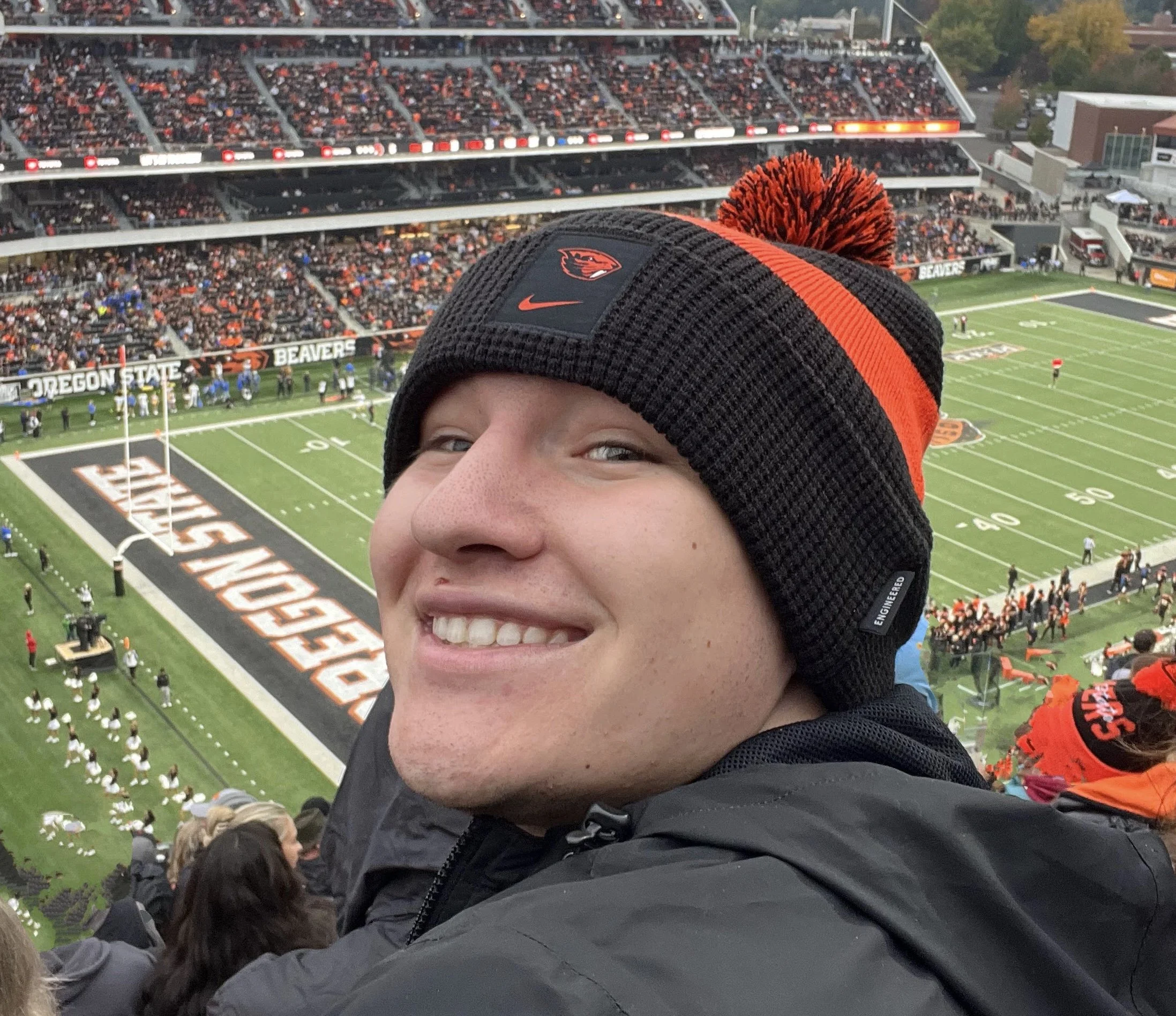 A person smiling at the camera while at a football game at Oregon State Stadium, with the field and crowds in the background.