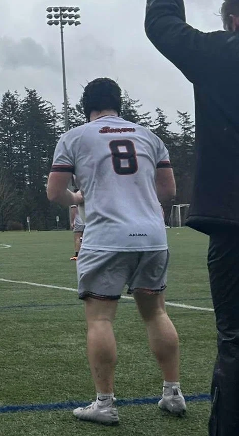 A soccer player wearing a white jersey with the number 8 on the back, gray shorts, and white cleats, standing on a soccer field with trees in the background.