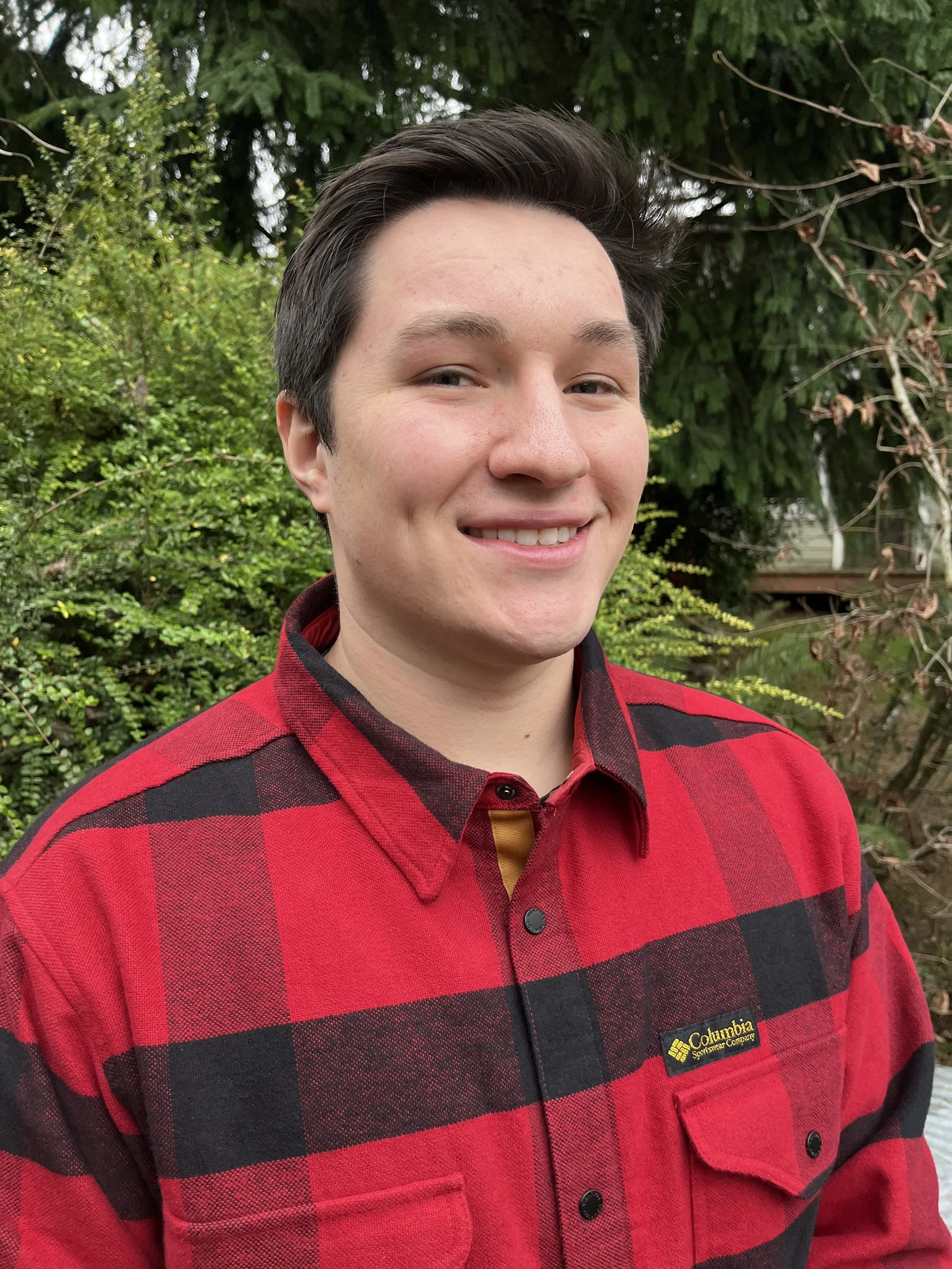 A young man smiling in an outdoor setting with green trees and bushes in the background. He is wearing a red and black plaid Columbia shirt.
