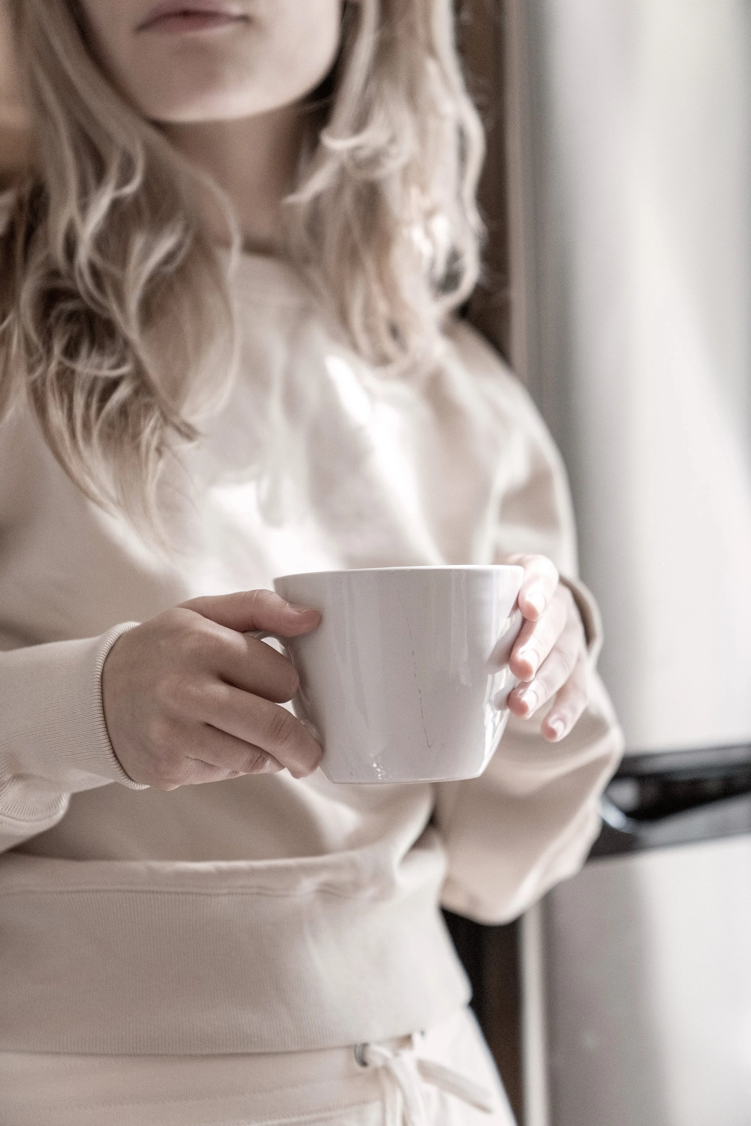 Person holding a white ceramic mug while standing indoors, with long, curly blonde hair, wearing a beige hoodie.
