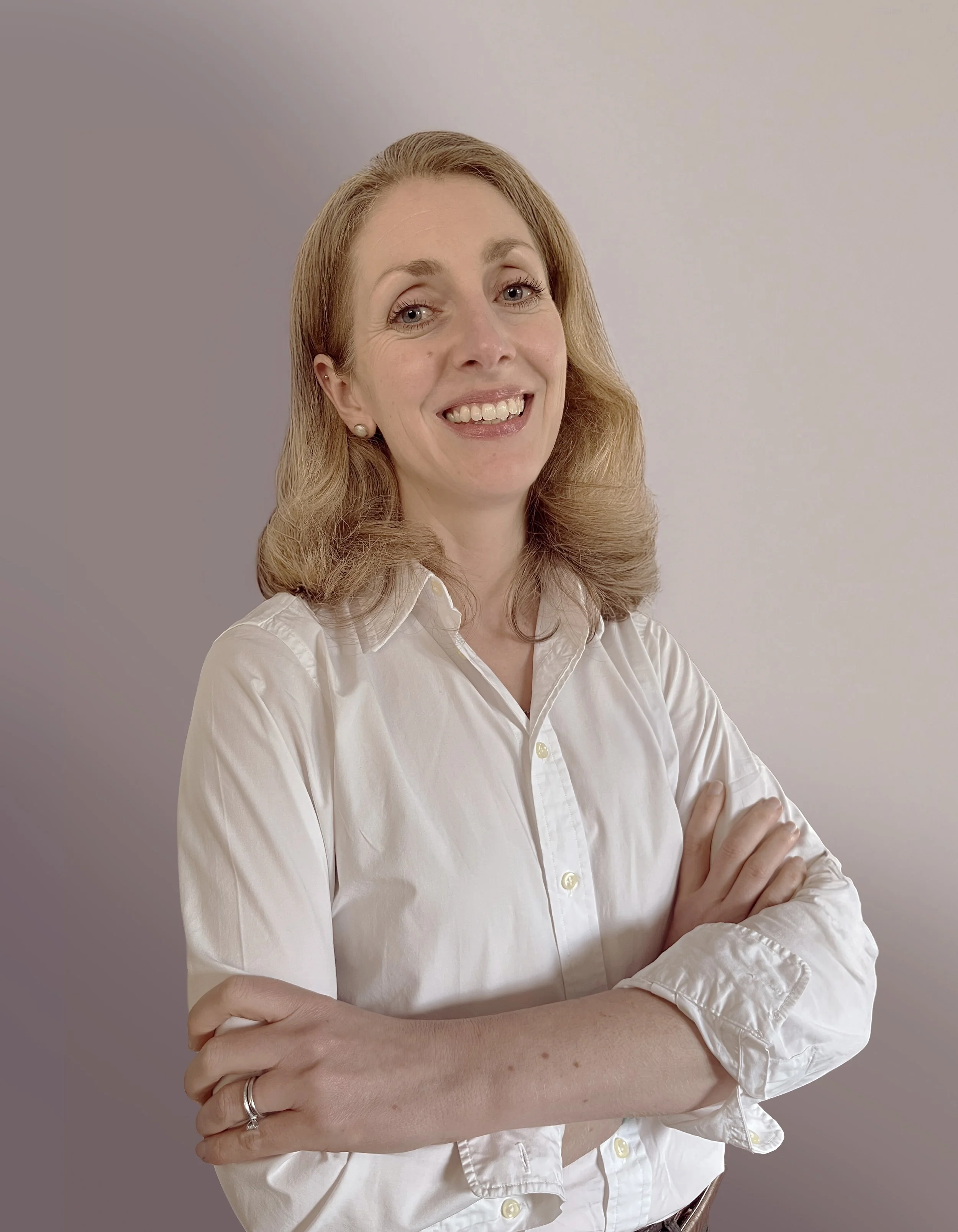 A woman with shoulder-length reddish hair wearing a white button-up shirt, smiling and crossing her arms, standing against a plain light-colored background.