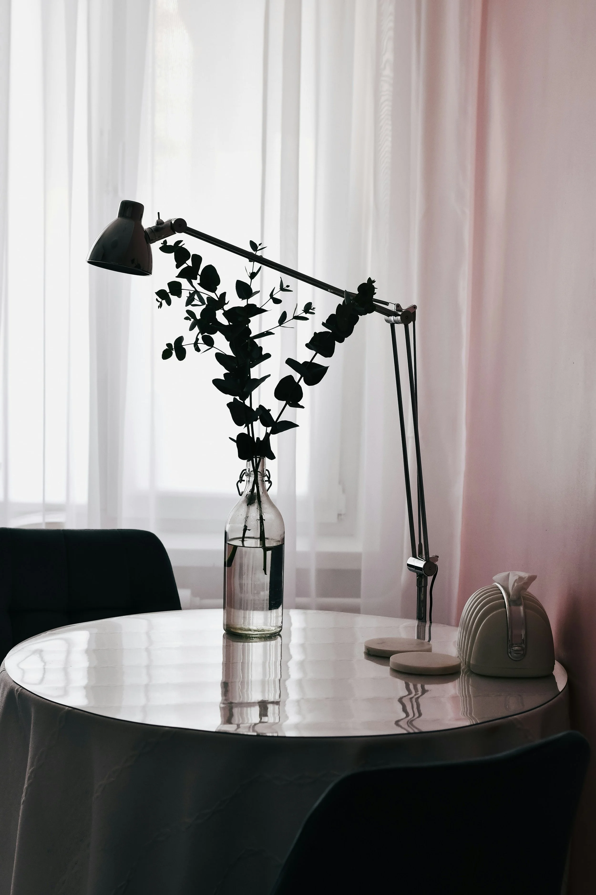 A round dining table with a glass vase holding a leafy branch, a white tissue box, and coasters, illuminated by backlit sheer curtains.