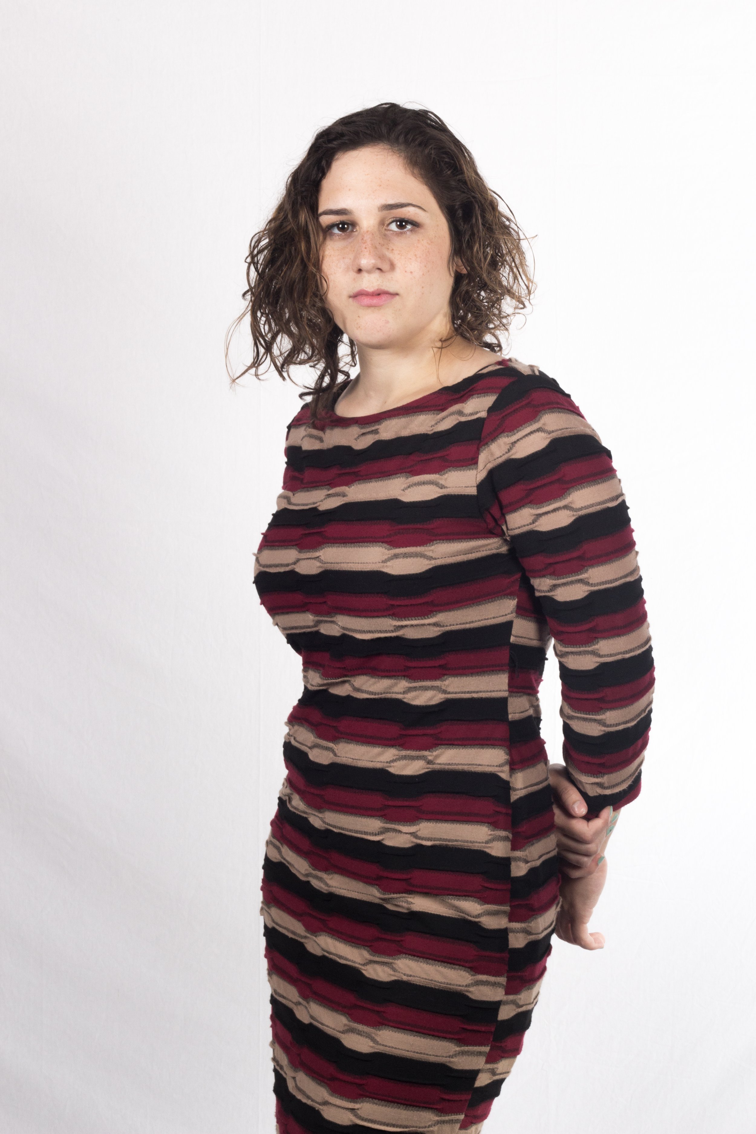 A young woman with shoulder-length curly brown hair, wearing a fitted, long-sleeve dress with horizontal stripes in beige, black, and red, standing against a plain white background, looking directly at the camera with a neutral expression.