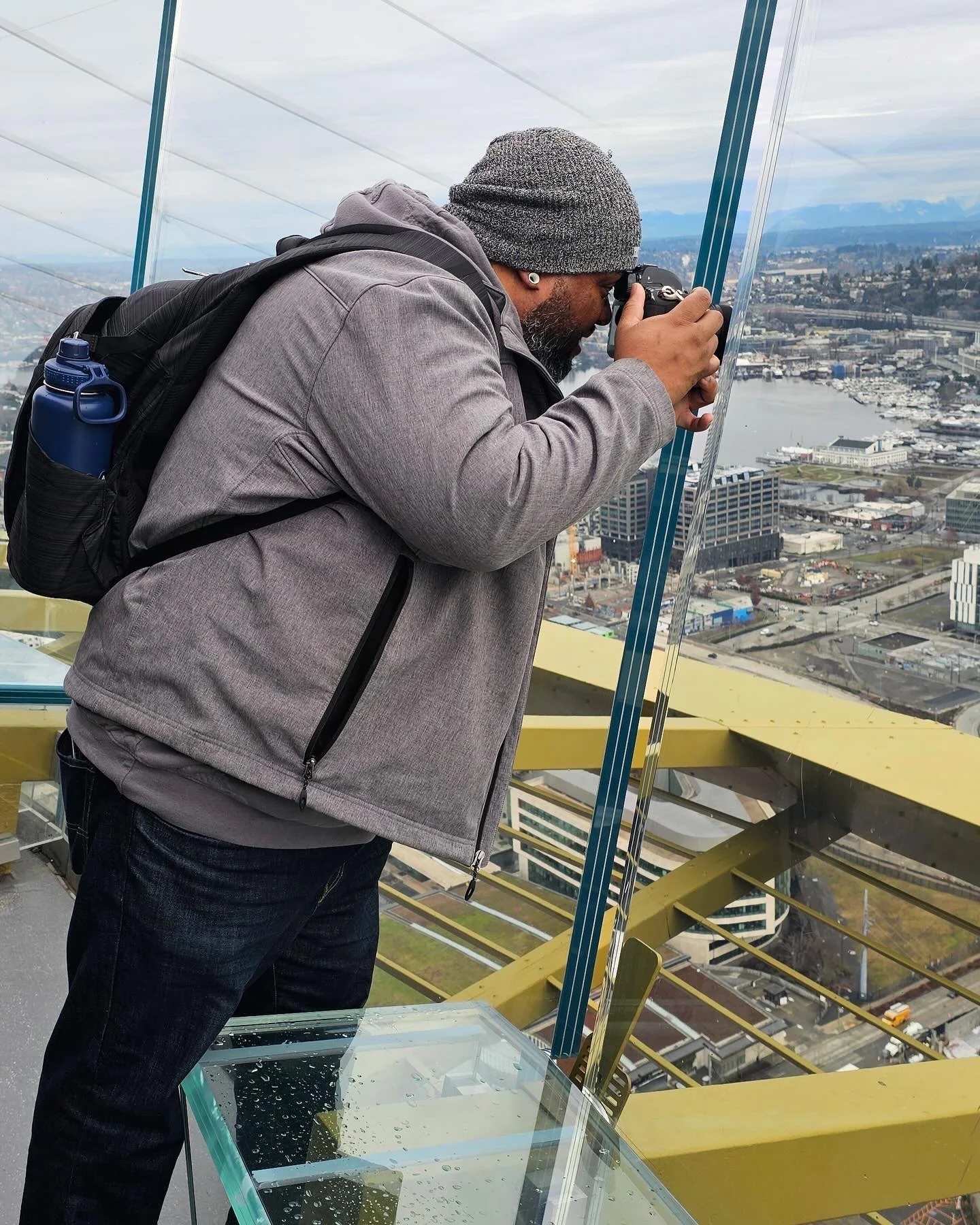 Man in gray jacket and gray beanie looking through a camera on an outdoor observation deck, cityscape and water in the background.