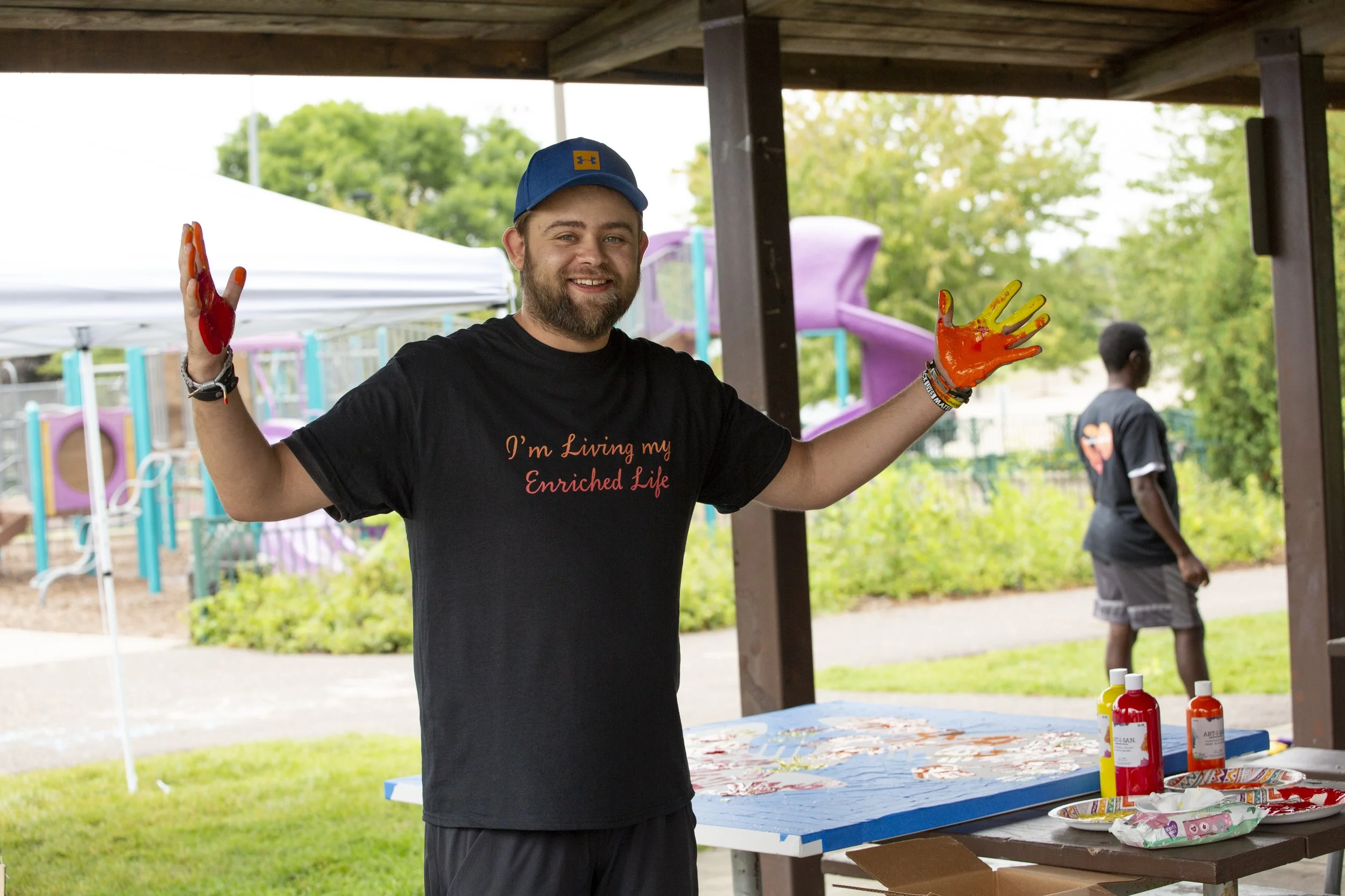 A smiling man with painted hands standing under a pavilion at an outdoor event, with a colorful playground in the background. He is wearing a black T-shirt with the words 'I'm Living my Enriched Life' and a blue cap. On the table in front of him are 