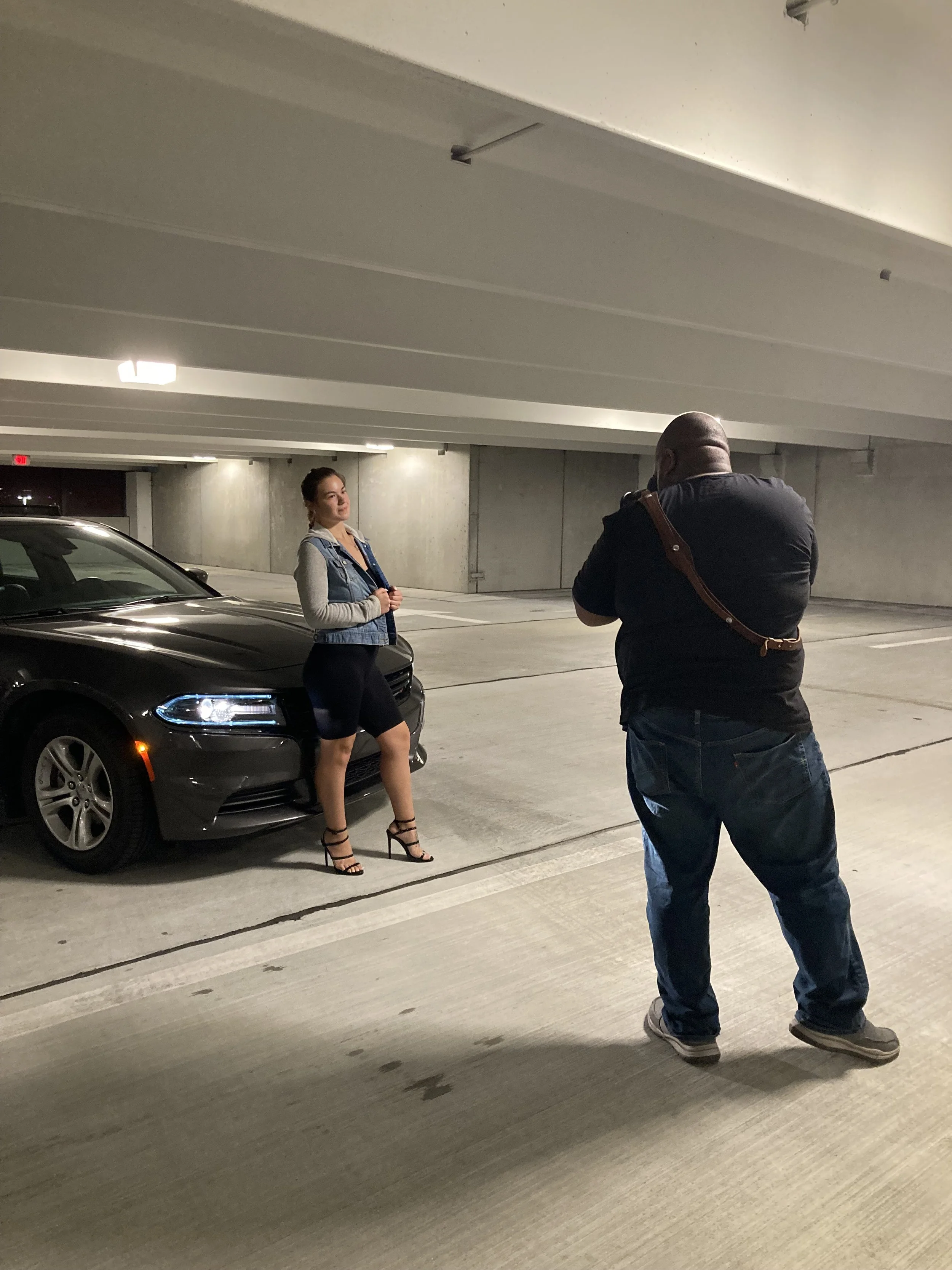 A woman in high heels and a denim vest posing for a photo with a man taking her picture in a parking garage.