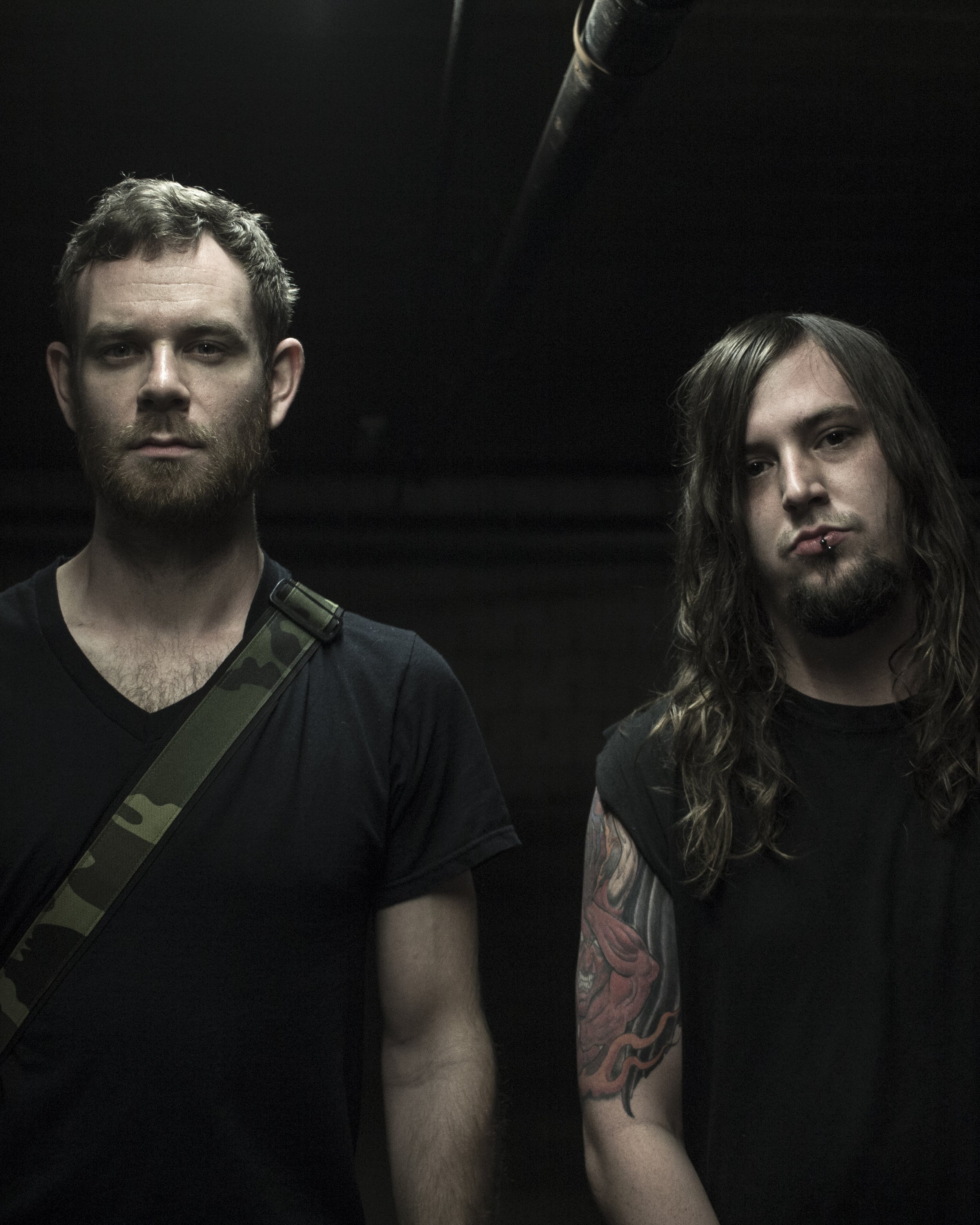 Two young men with long hair and beards, wearing black shirts, standing in a dark setting.
