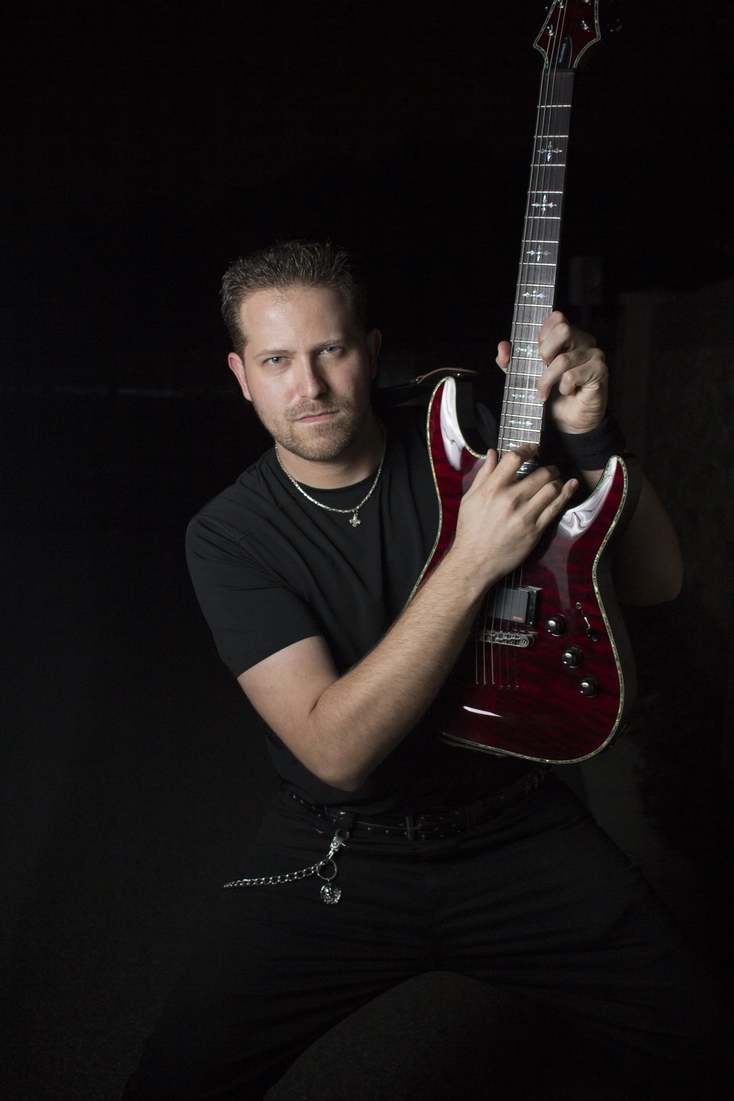A man with short hair and a beard, wearing a black t-shirt, holding a red electric guitar with a dark background.