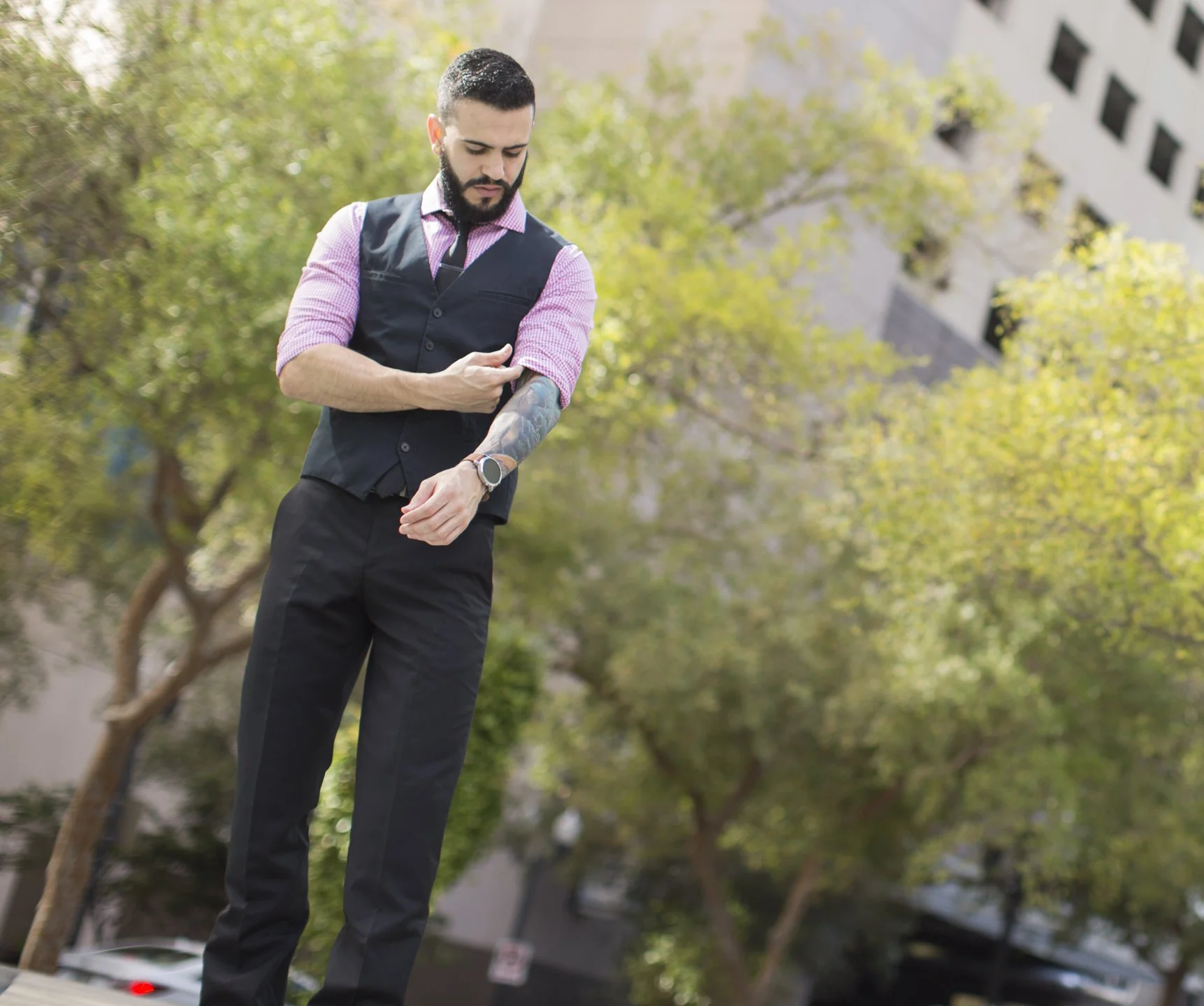 A man in a business vest and dress shirt checks his wristwatch outdoors with trees and a building in the background.