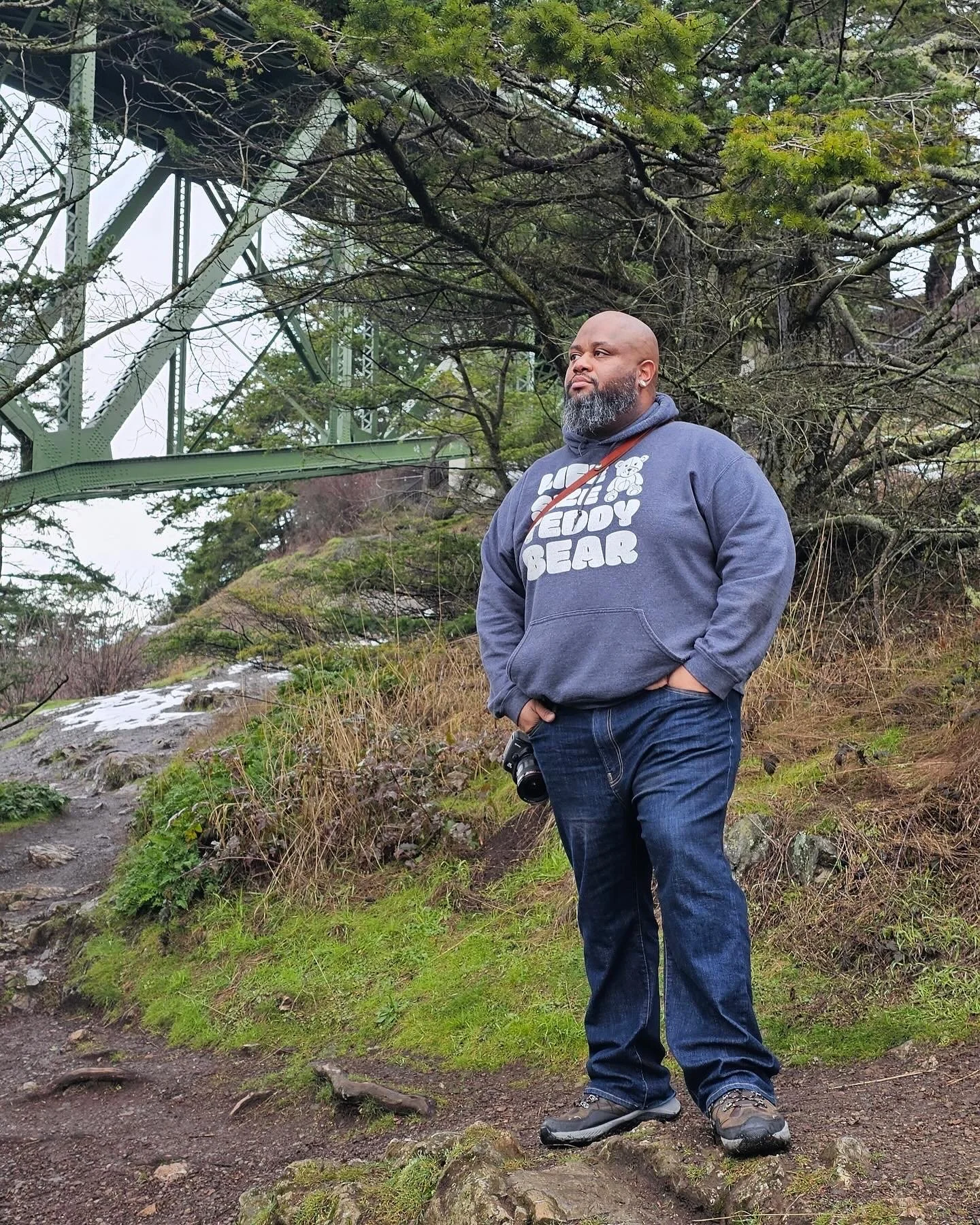 A man stands outdoors on a dirt path with moss and roots, with a camera hanging from his pocket. He's wearing a hoodie with the words 'UFC Teddy Bear' and a bear graphic, jeans, and hiking shoes. Behind him are trees, a mossy hillside, and a green bridge structure overhead.