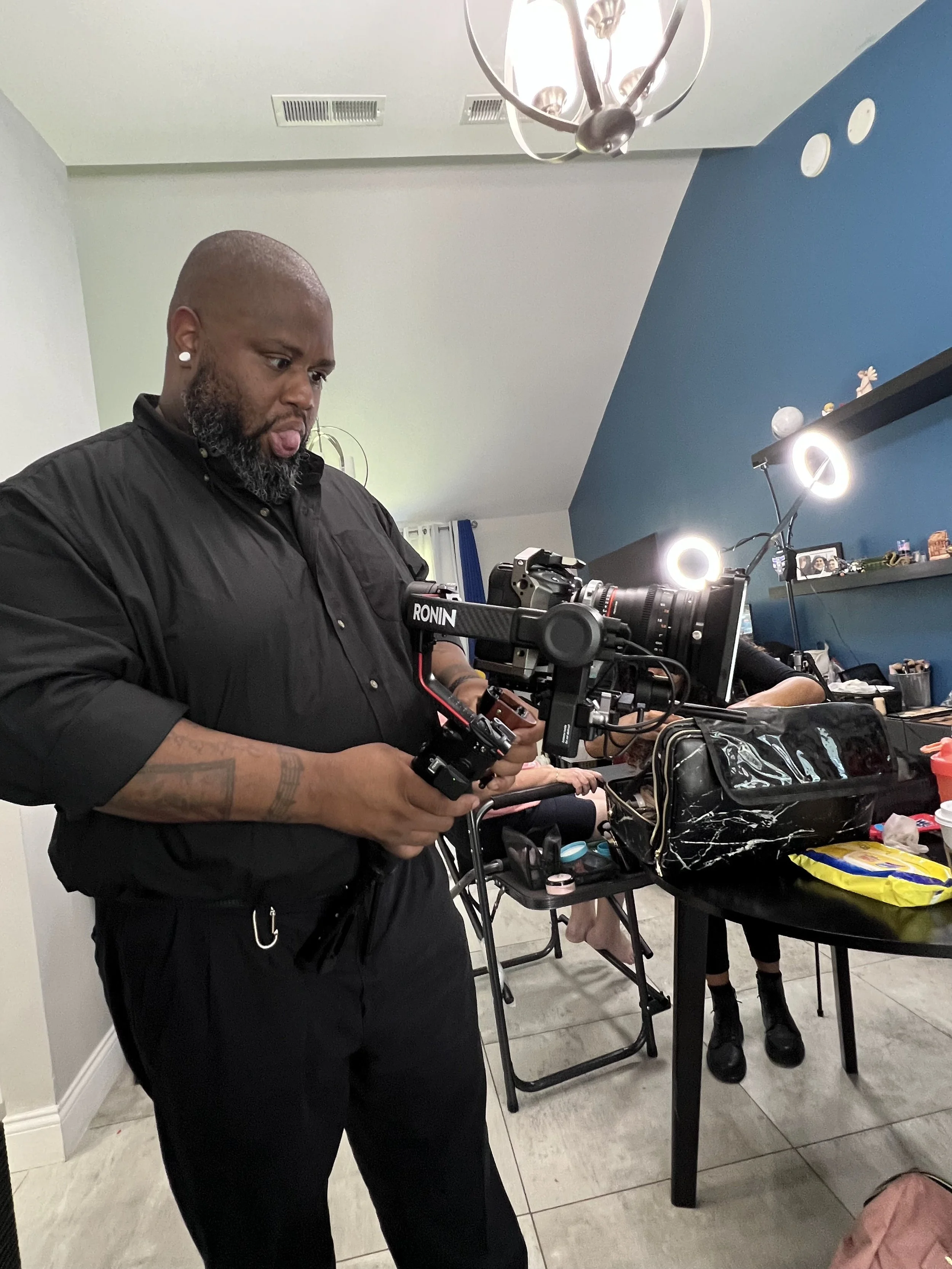 A man holding a camera stabilizer while filming indoors, with a table and various items in the background.