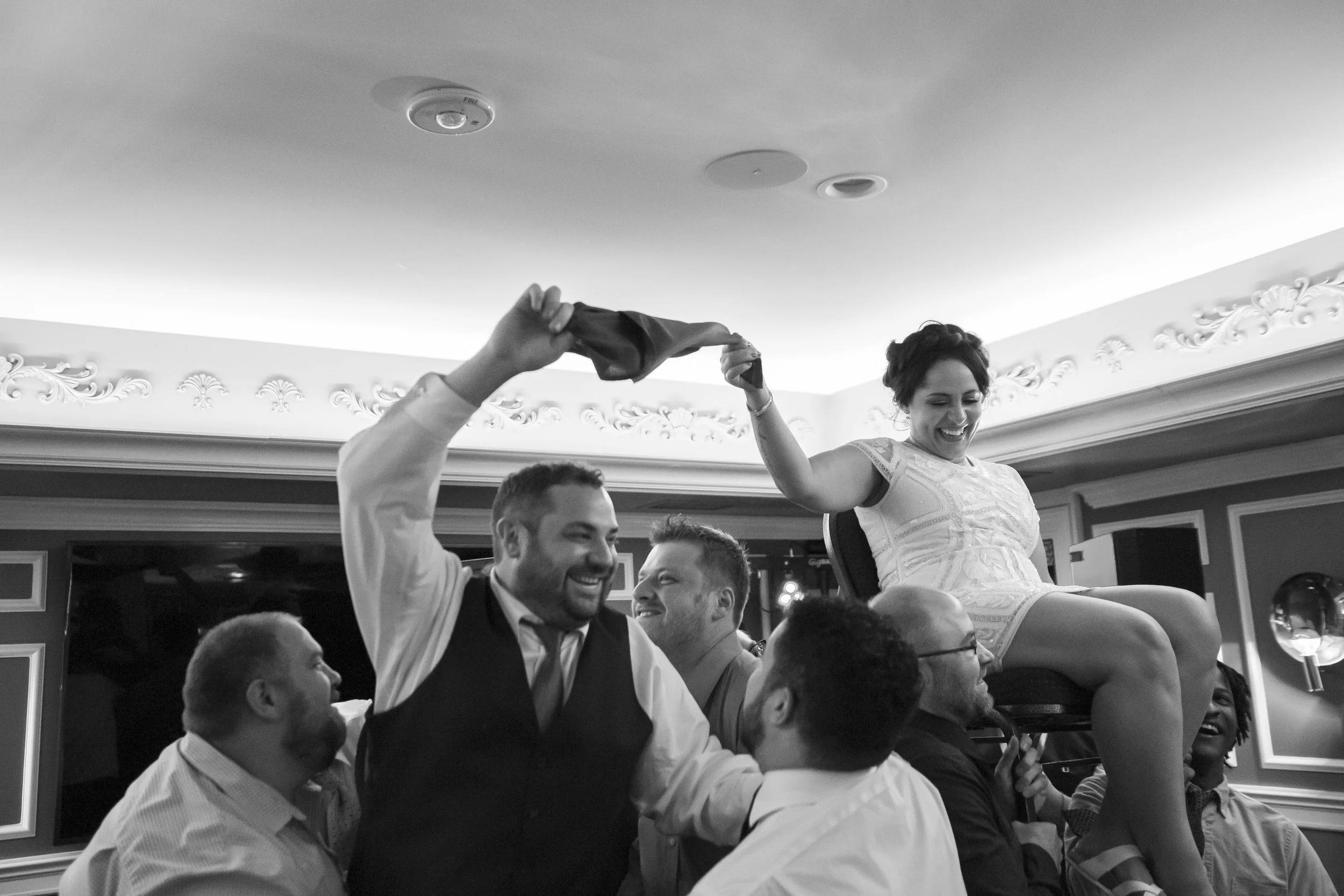 black and white photo of a couple holding a napkin together while being hoisted on chairs