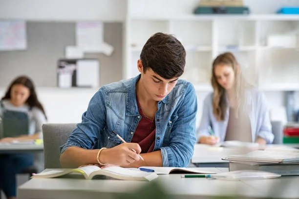 A young man sitting at a desk studying with notebooks and pens, in a classroom or library setting, with two other students in the background.