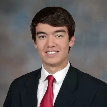 A young man with dark hair wearing a black suit, white shirt, and red tie, smiling in front of a neutral background.