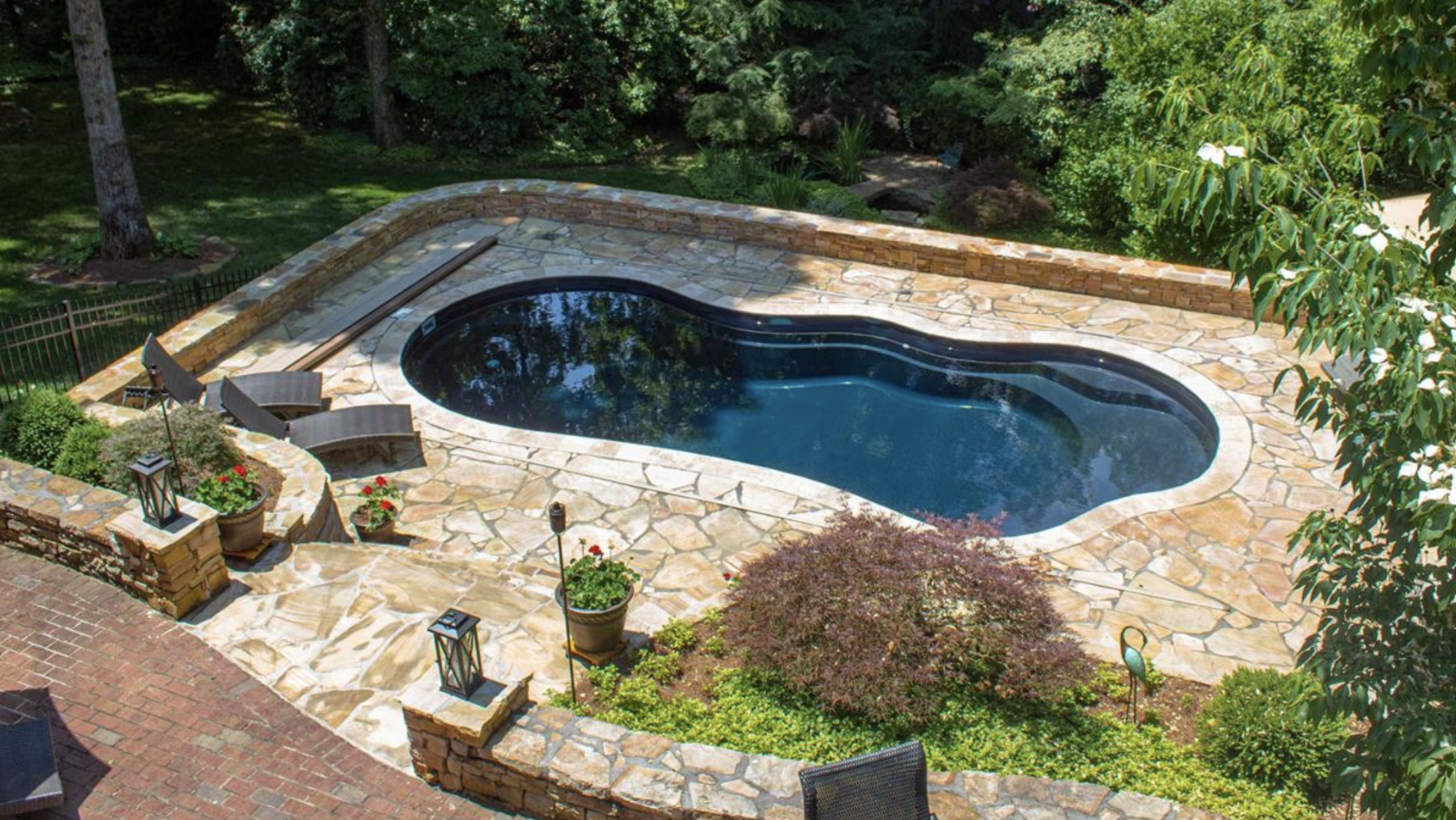 A backyard with a stone patio, a kidney-shaped swimming pool, surrounded by lush green trees and plants, with outdoor seating and lanterns.