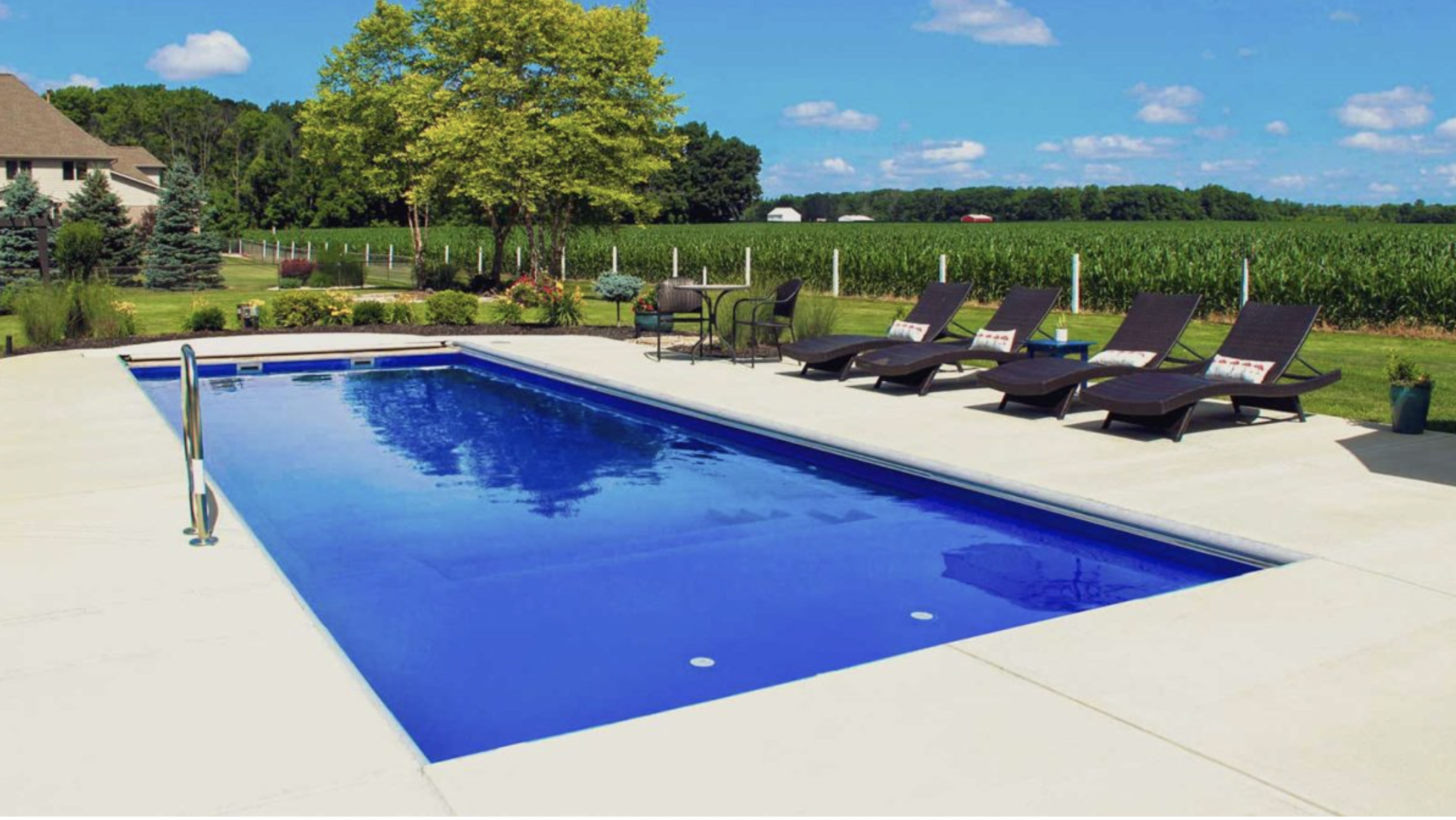 A backyard swimming pool with four lounge chairs, a small table, surrounded by a well-maintained garden and trees, with a green farm field and blue sky in the background.