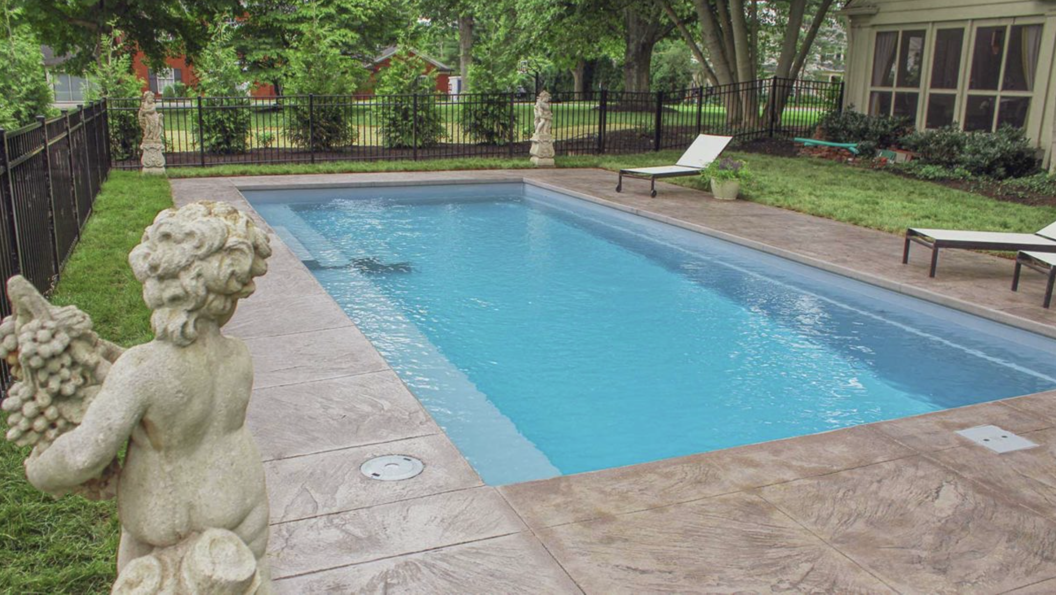 A backyard swimming pool surrounded by a paved deck, with decorative statues and lush green trees in the background.