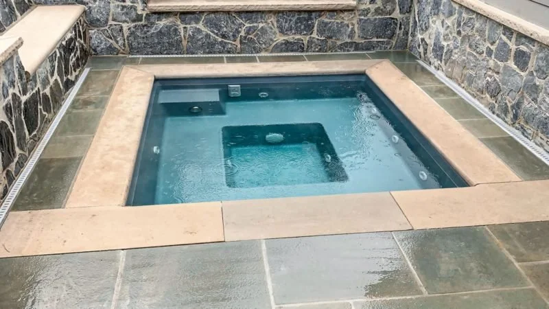 A small hot tub with built-in seating, surrounded by stone and tile patio, with some water bubbles on the surface.