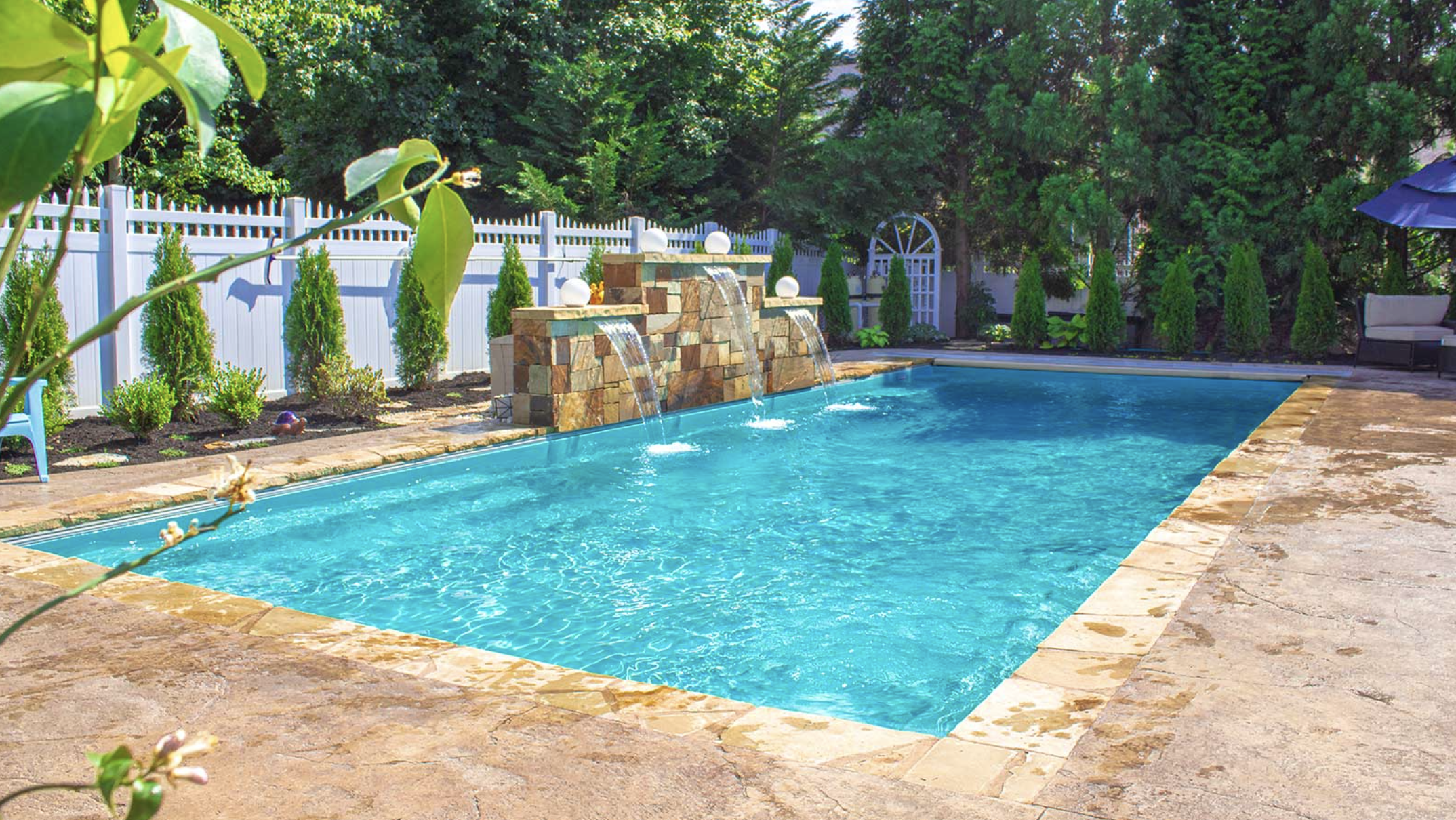 Backyard swimming pool with water fountain feature, surrounded by stone paving and green trees, with a white fence in the background.