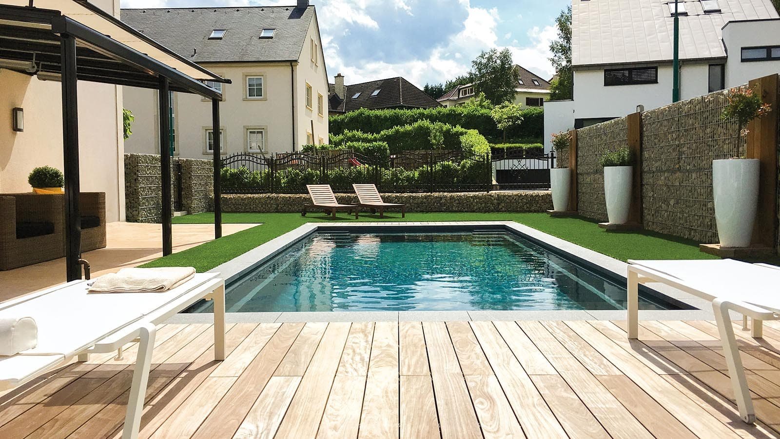 A backyard with a swimming pool, two lounge chairs, potted plants, and a patio area with outdoor furniture under a shaded pergola. The yard is enclosed by a fence and surrounded by residential houses.
