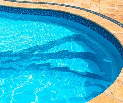 A section of a clear blue swimming pool with built-in steps and a surrounding tan stone deck.