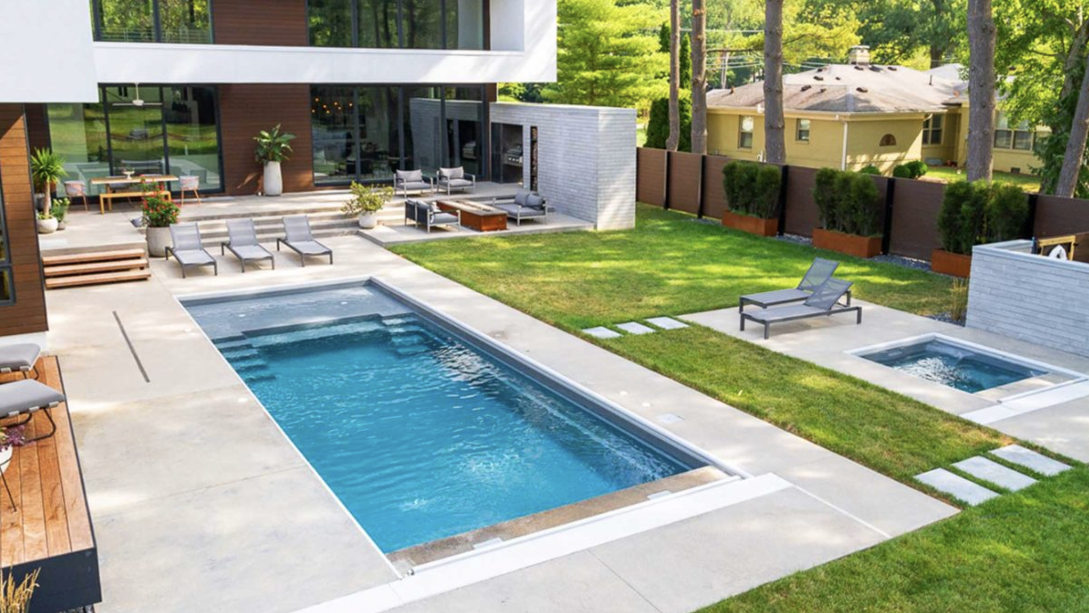 Modern backyard with a swimming pool, hot tub, and lounge chairs on concrete patio, surrounded by a green lawn and tall trees.