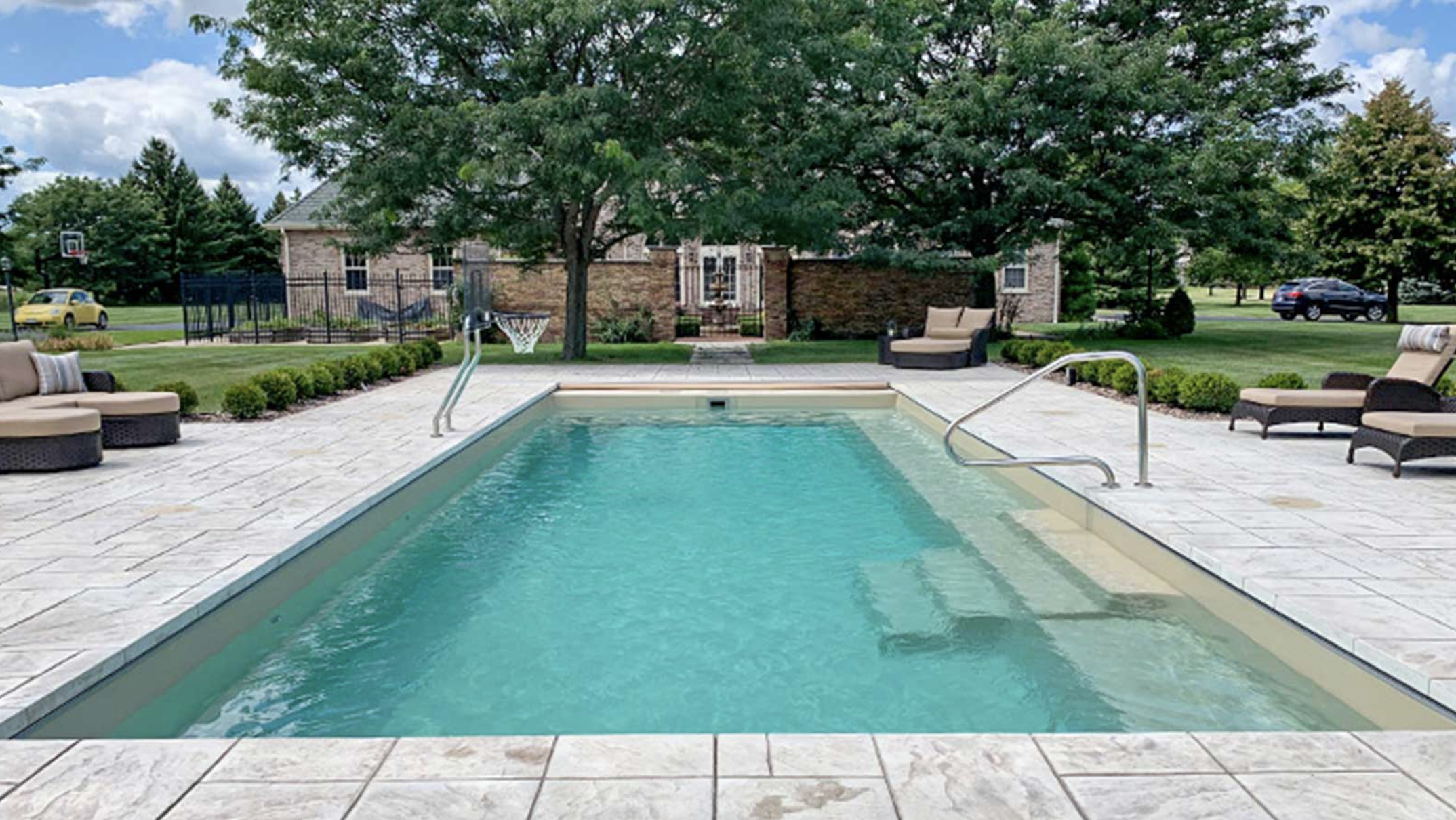 Residential backyard with swimming pool, surrounded by outdoor furniture, trees, and greenery, with a brick house and parked cars in the background.