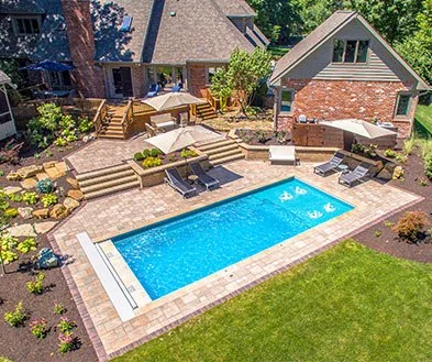 A backyard with a swimming pool, lounge chairs, and a wooden deck with outdoor furniture, surrounded by a landscaped garden and a house in the background.