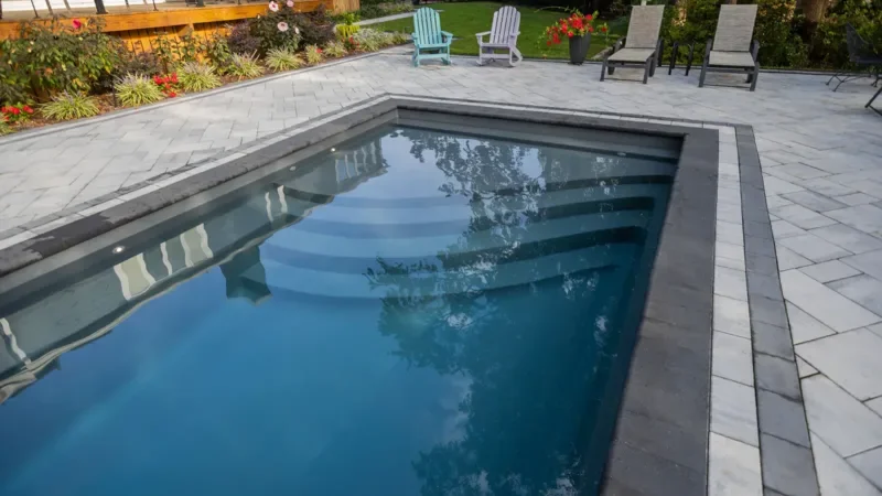 Backyard with a rectangular swimming pool, gray stone patio, and colorful Adirondack chairs, flower bed, and plants in the background.