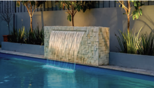 Water features a cascading waterfall over a stone wall into a swimming pool at night.