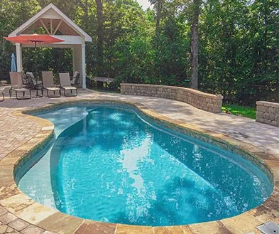 A backyard swimming pool with a stone patio surrounding it, shaded lounge chairs under a pavilion, and trees in the background.