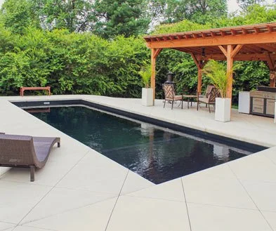 Backyard with a black rectangular swimming pool, outdoor furniture including a lounge chair, a shaded seating area with a wooden pergola, and lush green trees and bushes in the background.