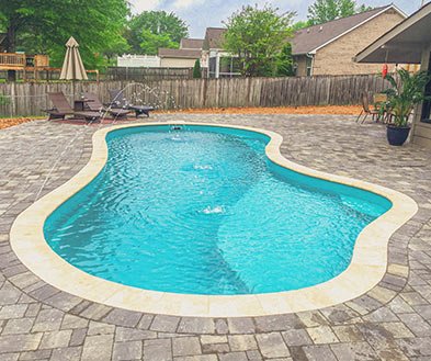 Backyard swimming pool with a curved shape, surrounded by gray paving stones, with a wooden fence, outdoor furniture, and trees in the background.