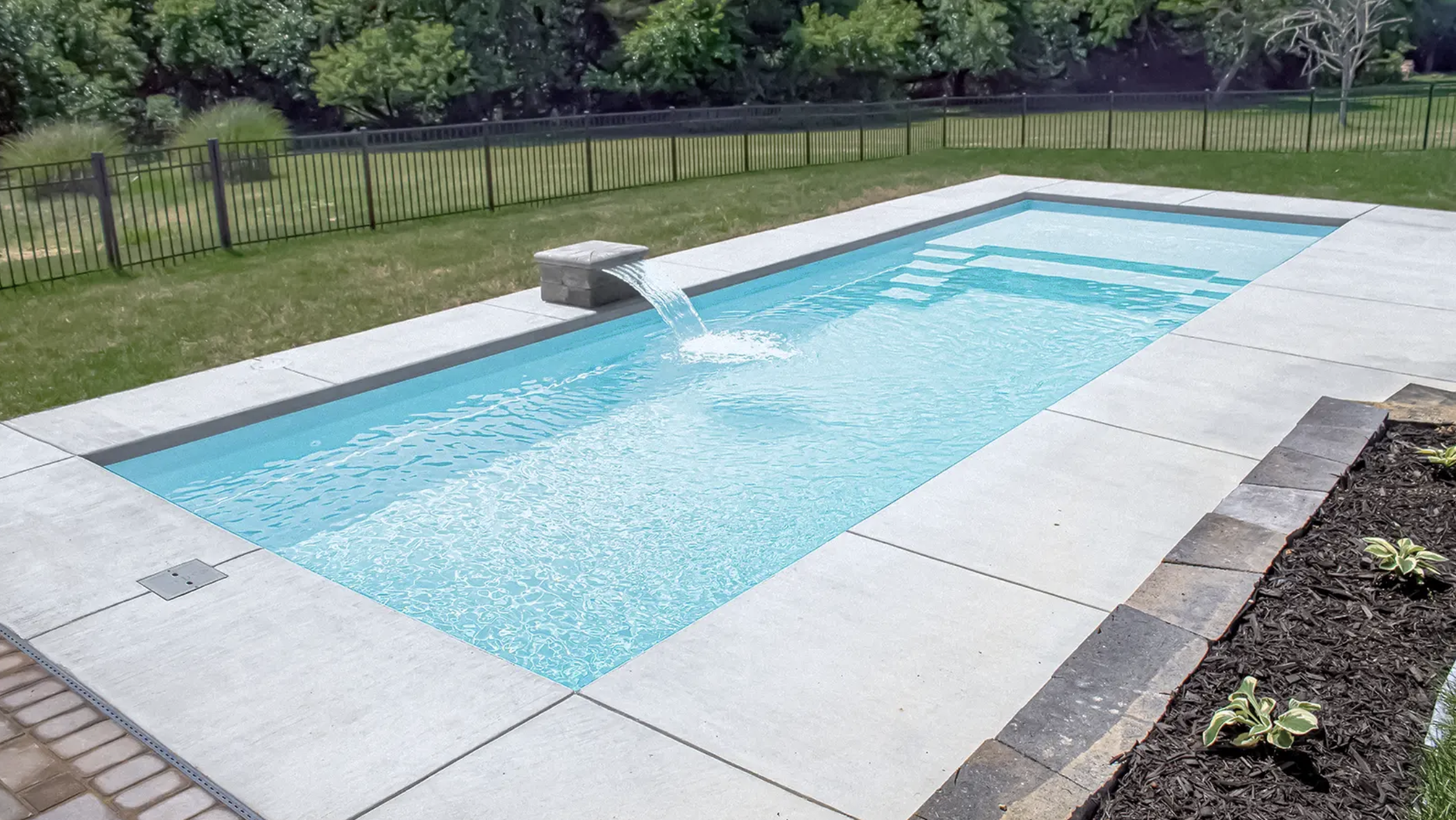 A rectangular backyard swimming pool with a waterfall feature on one end, surrounded by light-colored stone decking and a black metal fence, with lush green trees and grass in the background.