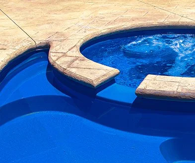 Close-up of a pool with a built-in hot tub or spa, featuring blue water and beige stone tile surrounding the area.