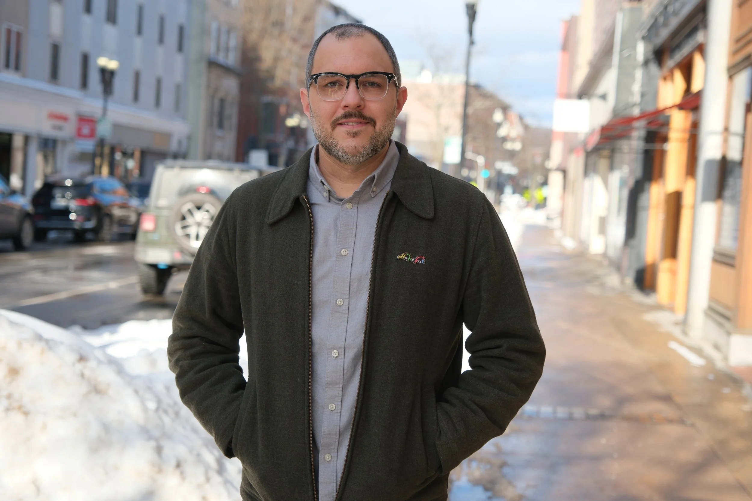 Scott Harriman standing on a sidewalk in a city street during winter, wearing glasses and a dark jacket, with snow on the ground and parked cars in the background.
