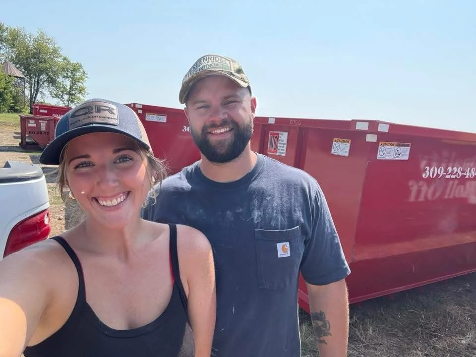 A smiling woman and man taking a selfie outdoors, with red dumpsters and trees in the background on a sunny day.