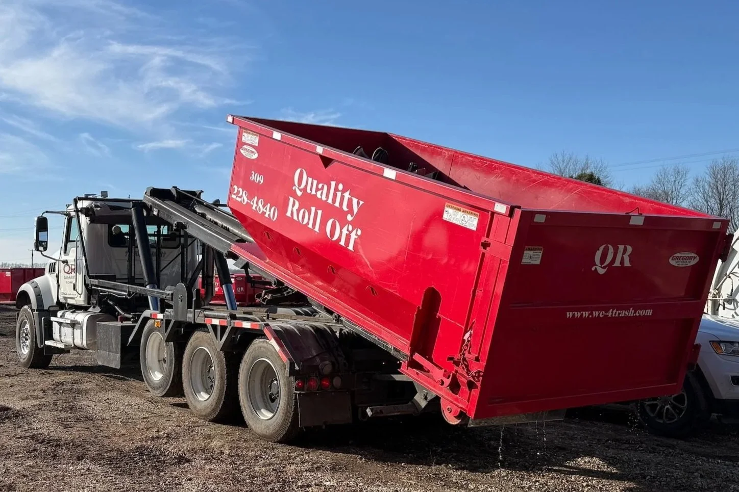 A red dump truck with a tilted bed, displaying the words "Quality Roll Off" on the side, parked on a gravel lot under a clear blue sky.