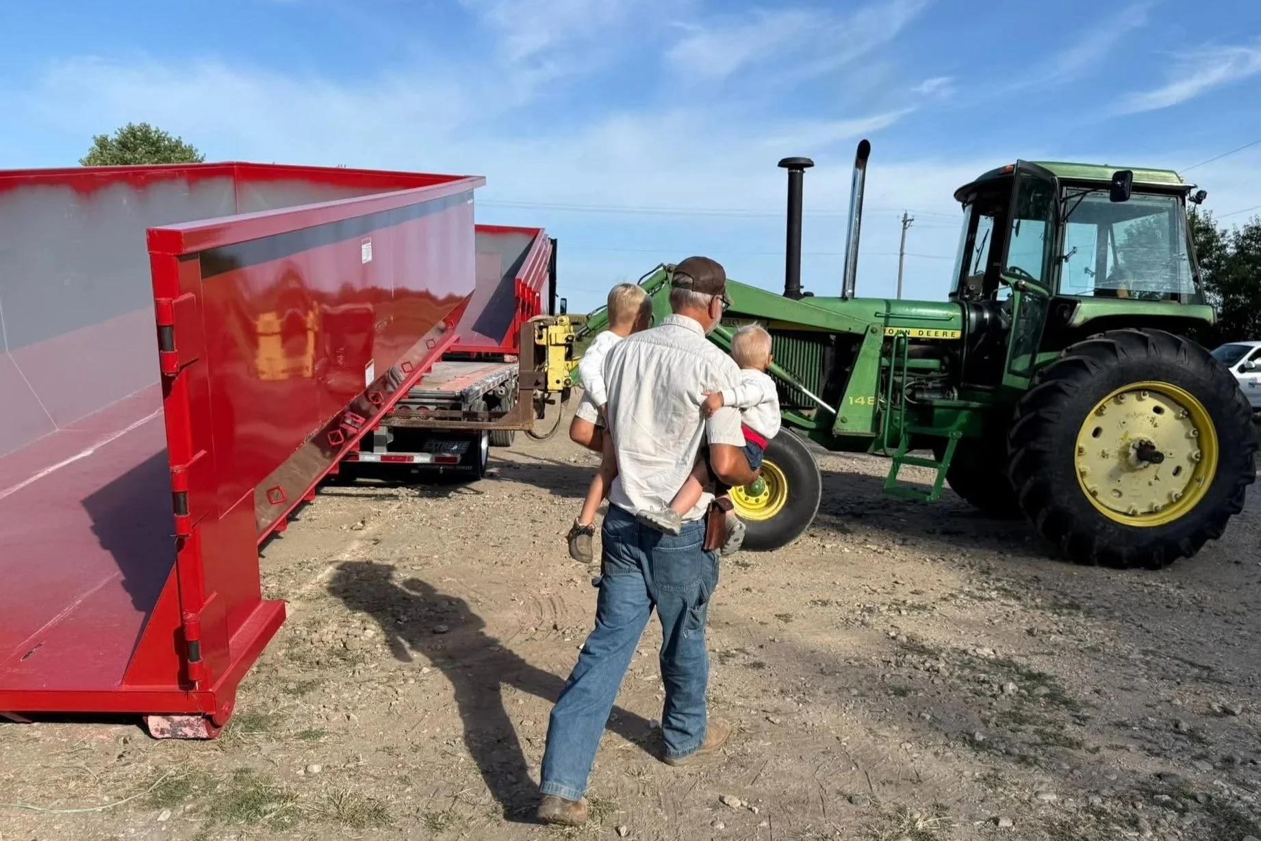A man holding two children while standing near a green John Deere tractor attached to a red trailer on a dirt lot under a blue sky.