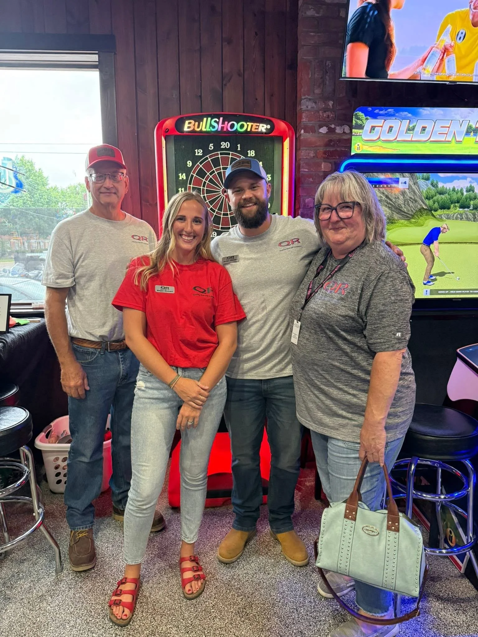 Group of four people standing together indoors, smiling for the photo, with an electronic dartboard and a video game screen showing a golf simulation behind them.