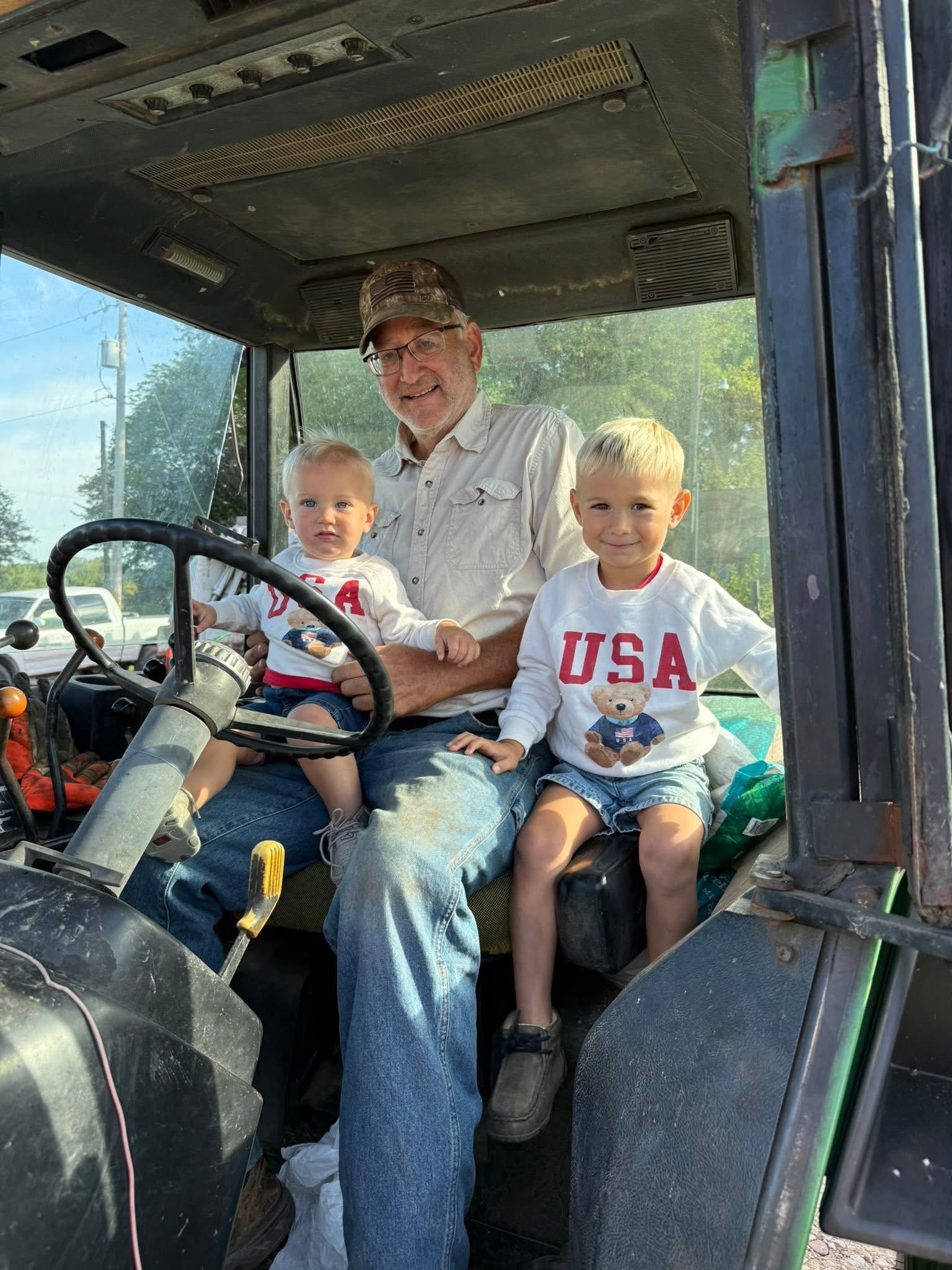 A smiling man with glasses and a camouflage cap sitting in the driver’s seat of a tractor with two young children, both wearing USA shirts, inside a farm vehicle on a sunny day.